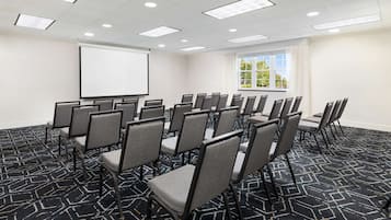 Ein Konferenzraum mit Reihen von Stühlen, einer Projektionsleinwand und einem großen Fenster mit Vorhängen.