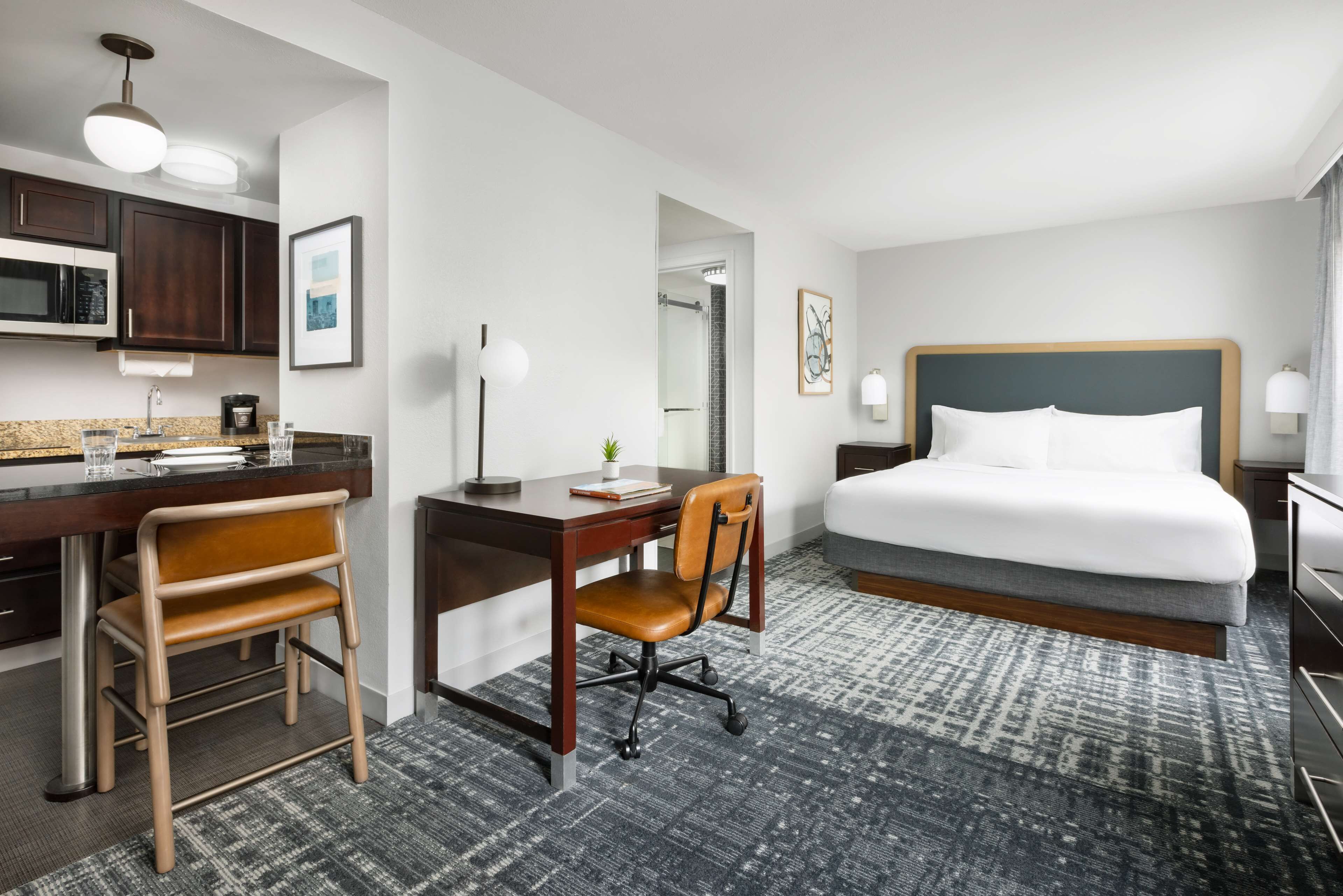 A hotel room with a bed, desk, chair, and kitchenette.