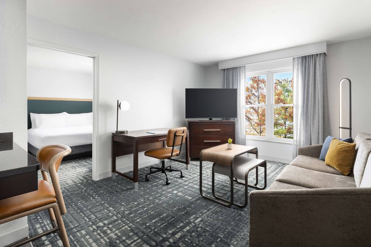 A hotel room with a bed, desk, chair, TV, sofa, and a window with a view of trees.