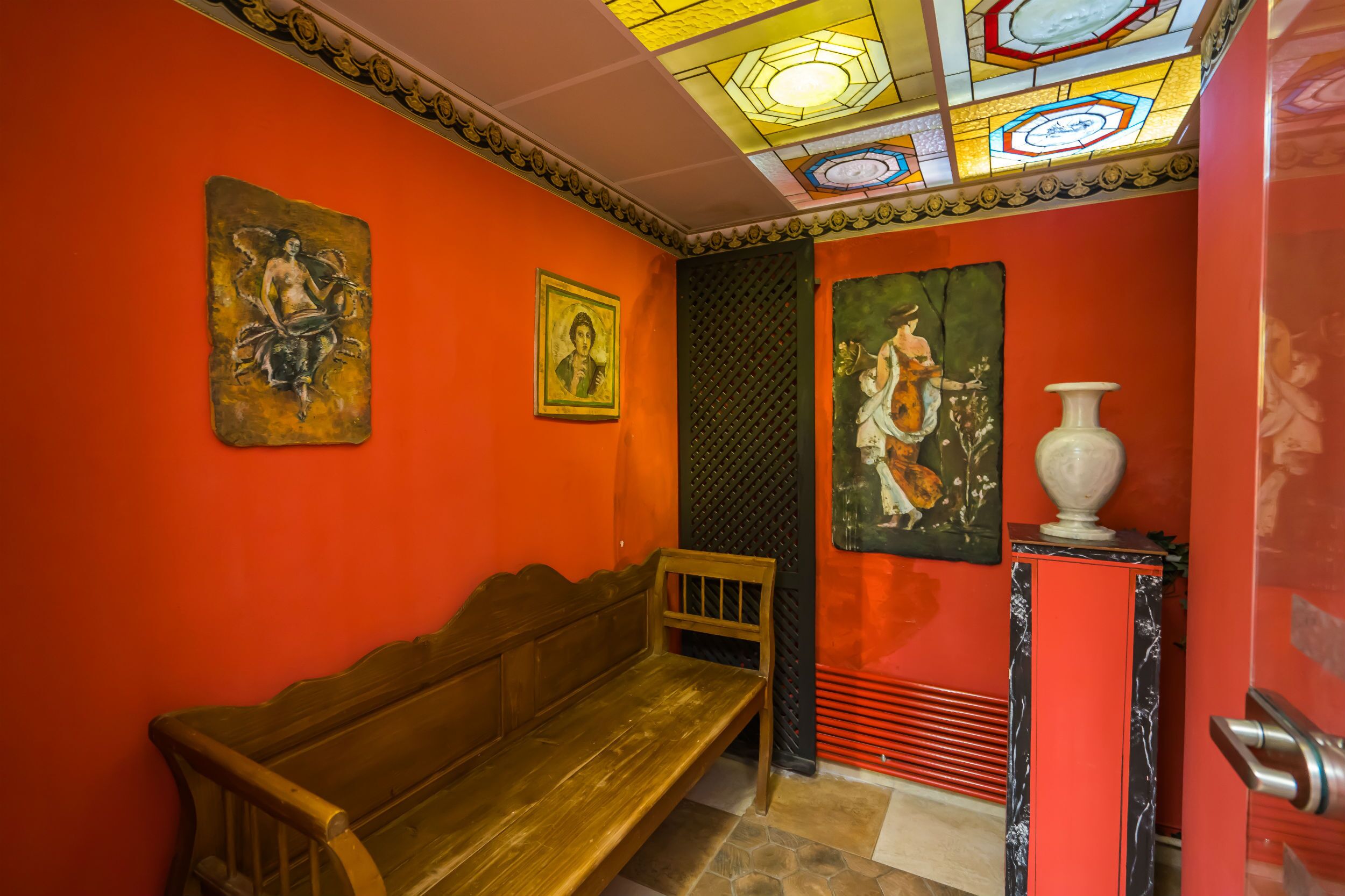 A room with a wooden bench, a vase on a pedestal, and artwork on the walls.