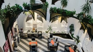 An indoor dining area with checkered flooring, tables with orange and black cushions, and potted plants. - Janus Boutique Hotel & Spa (Siófok)