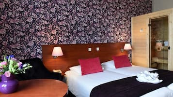 Hotelkamer met bloemenbehang, een houten hoofdbord, twee bedlampen, een bed met wit en rode beddengoed, een kleine ronde tafel met een paarse vaas, en een houten deur die leidt naar een badkamer.