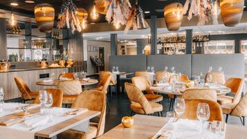 Un interior de restaurante con mesas redondas, sillas acolchadas, lámparas colgantes y una zona de bar al fondo.