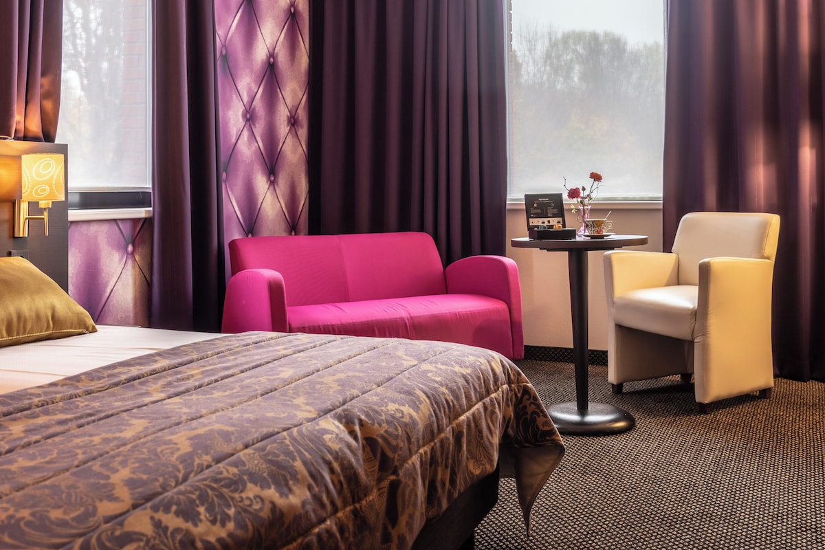 A hotel room with a large bed, a pink sofa, a white armchair, and a small table with a radio.