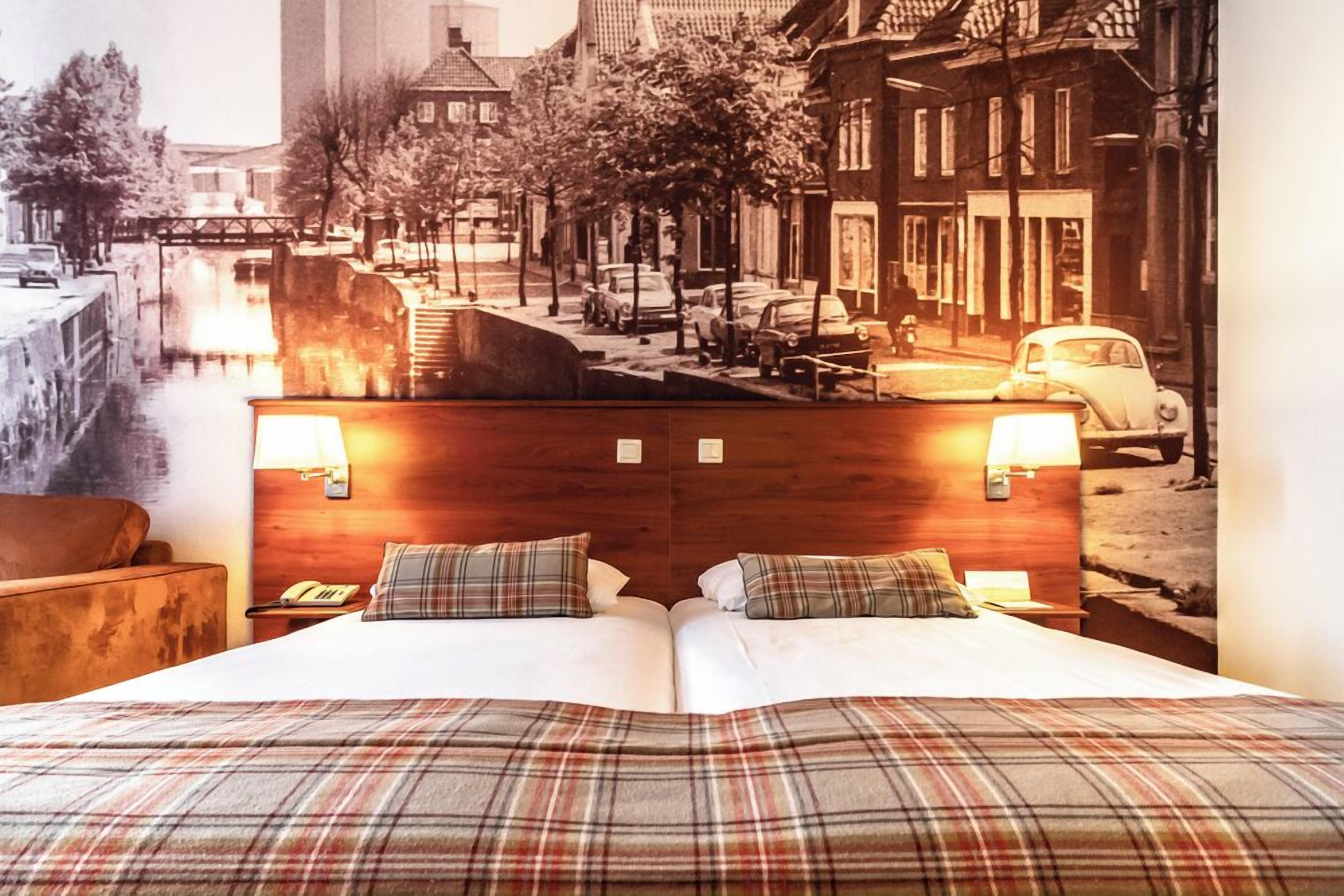 A hotel room with a large bed, two bedside lamps, a wooden headboard, and a cityscape mural on the wall.