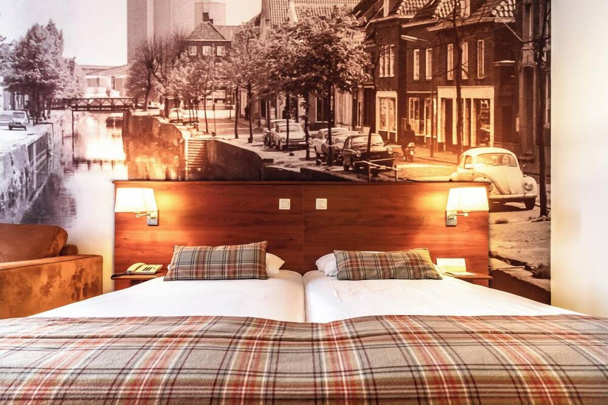 Habitación de hotel con una cama grande, dos lámparas de noche, un cabecero de madera y un mural con vistas a la ciudad.