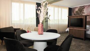 Habitación de hotel con una mesa de comedor redonda, sillas tapizadas en negro, un jarrón con flores secas, un televisor de pantalla plana y un cuadro en la pared.