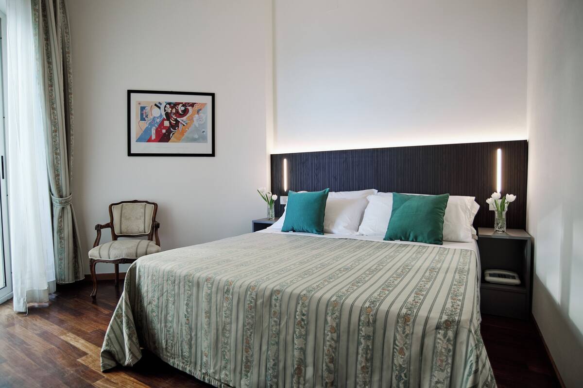 A neatly arranged bedroom with a large bed, a chair, a nightstand with a lamp, and a framed picture on the wall.