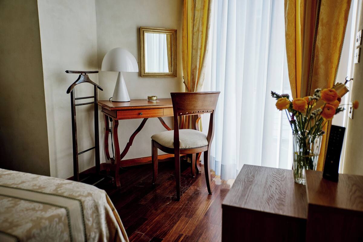 A hotel room with a wooden desk, a chair, a lamp, a mirror, and a vase with flowers.
