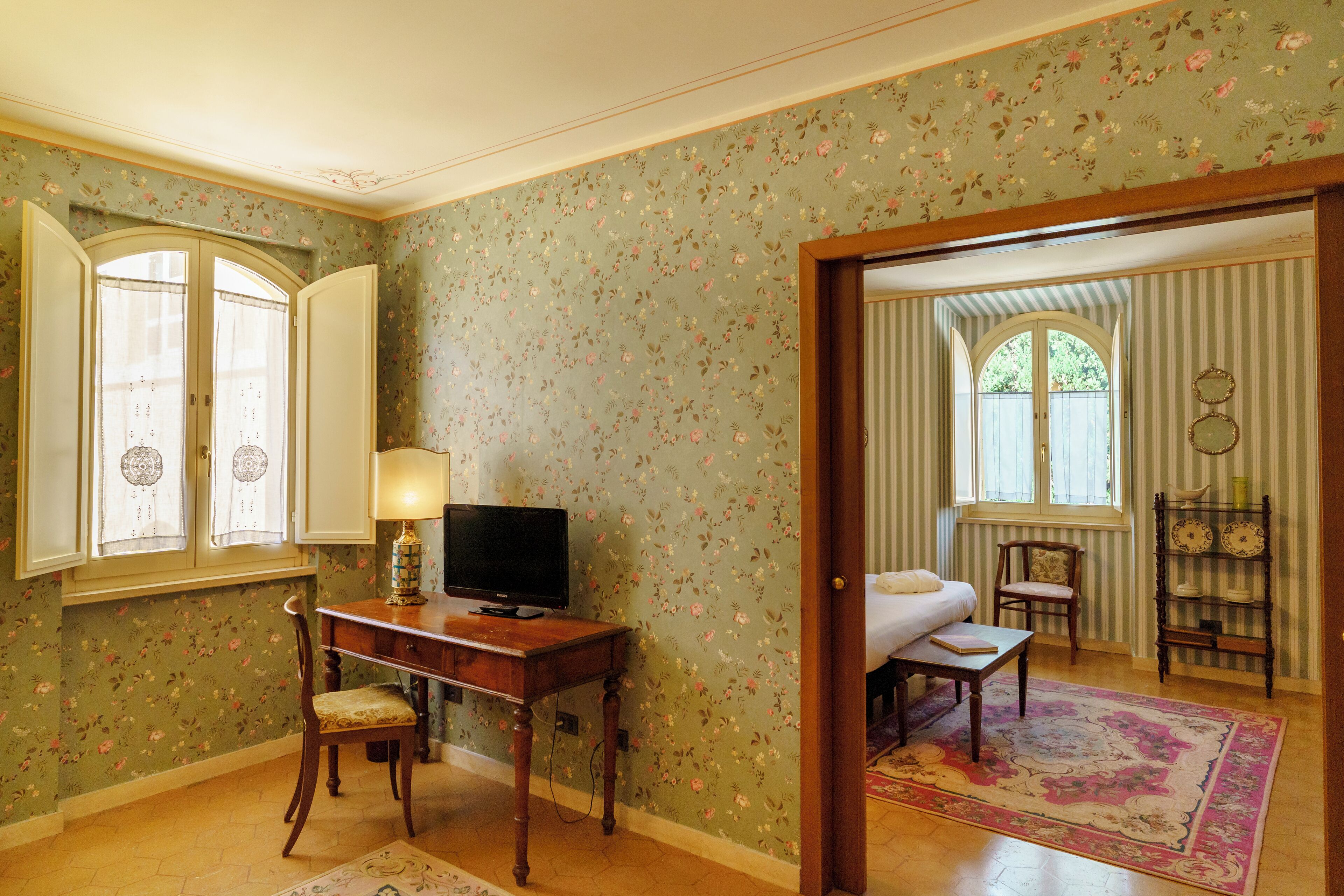 A room with floral wallpaper, a desk with a lamp, a television, and a bed with a view of the outdoors.