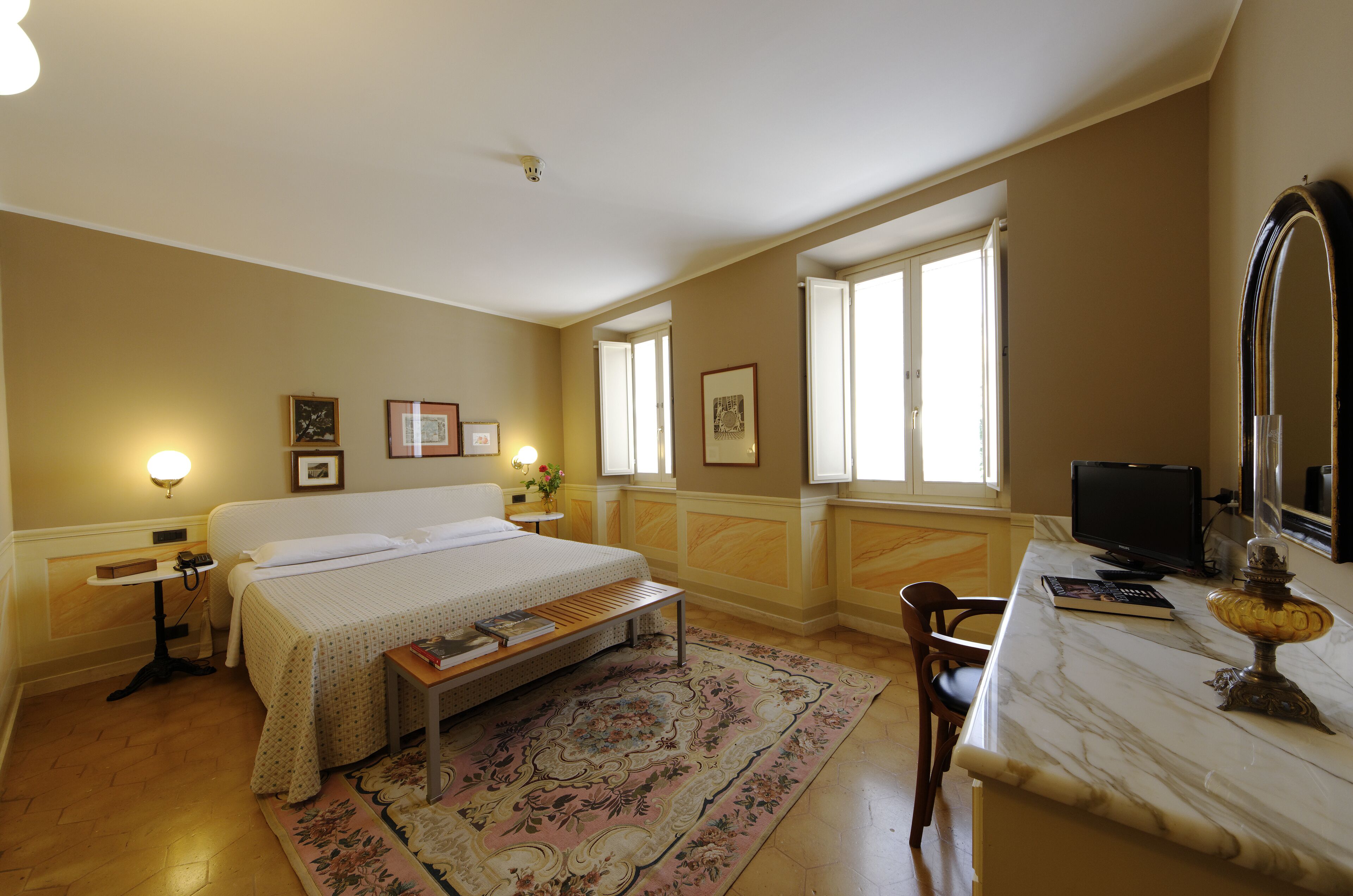 Ein Hotelzimmer mit Bett, Nachttisch, Lampe, Stuhl, Schreibtisch, Computer und einem großen Spiegel.