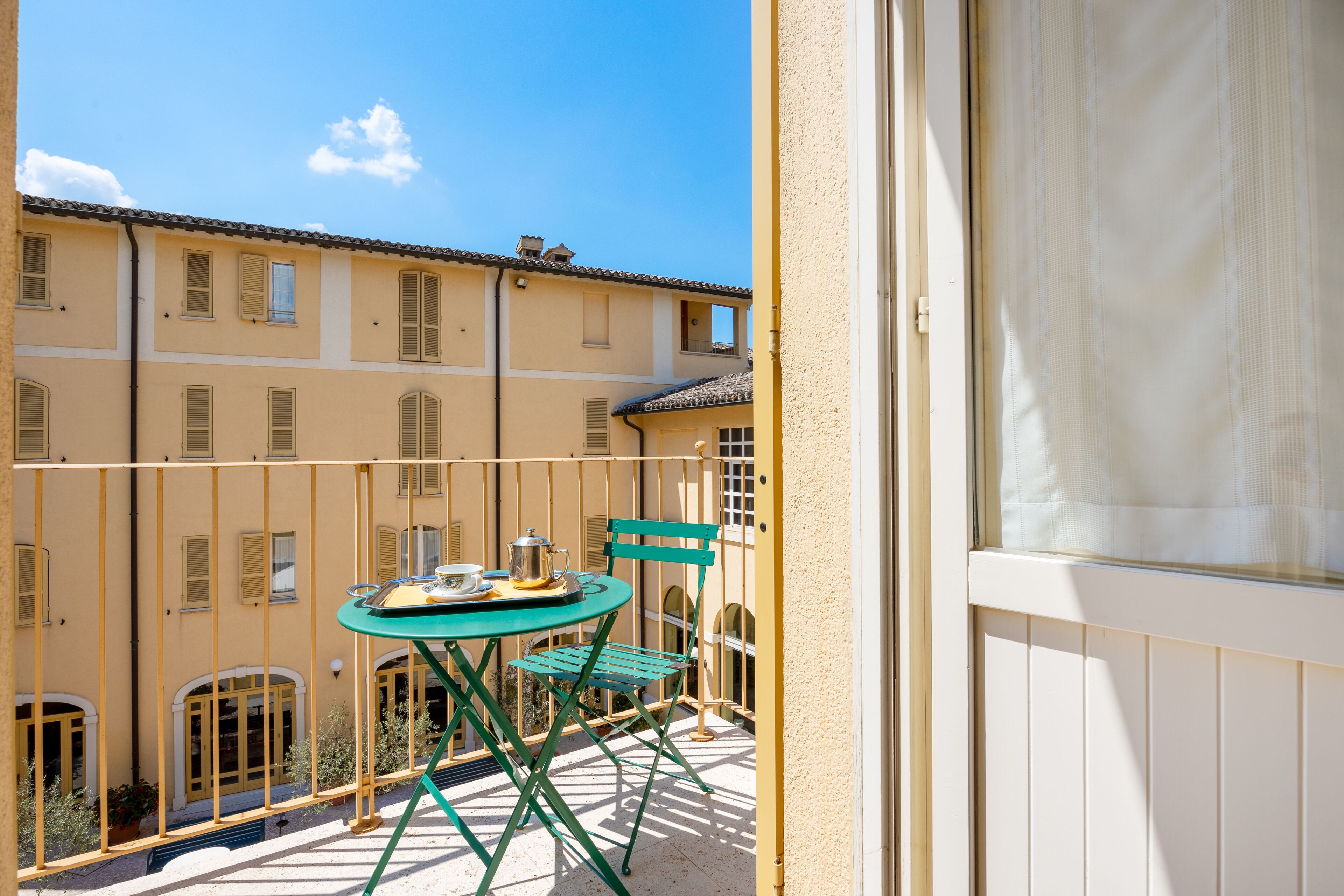 A balcony with a table and chairs, a view of a building with multiple windows, and a clear sky.