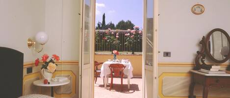 A hotel room with a bed, a bedside table with a vase of flowers, a chair, a table with a tablecloth, and a balcony with a table and chairs.