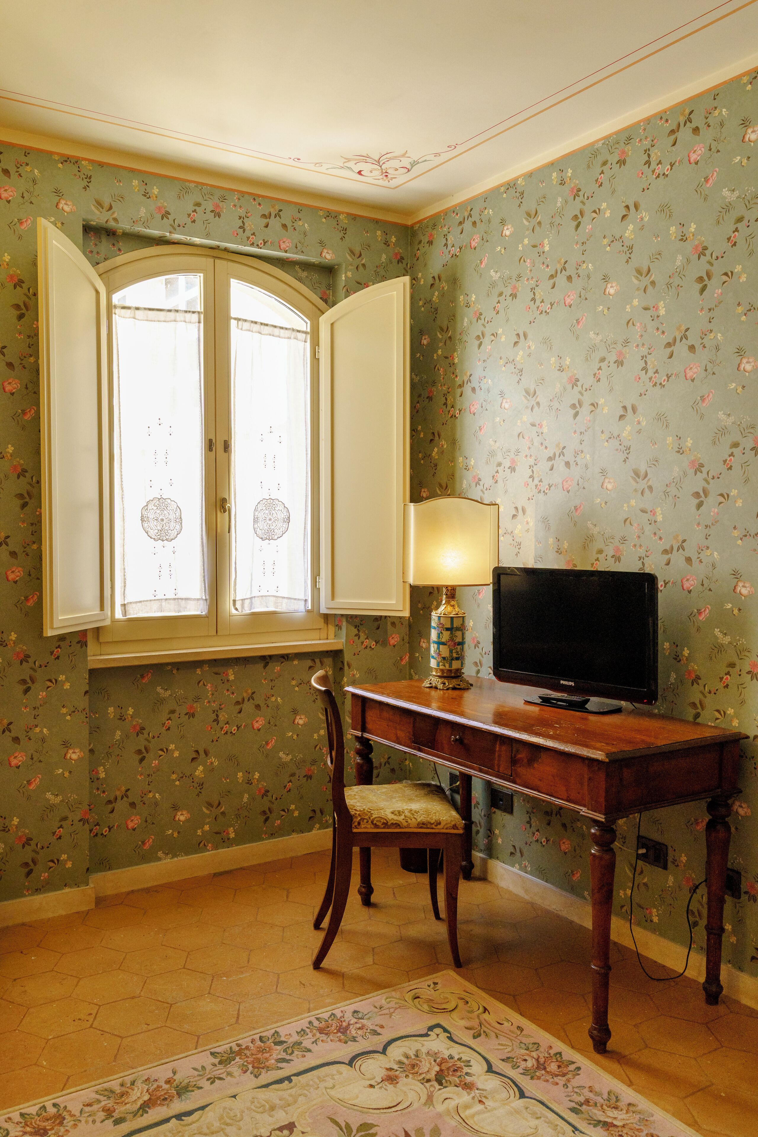 Habitació amb paper pintat floral, una taula de fusta amb televisió, una cadira i una finestra amb cortines.