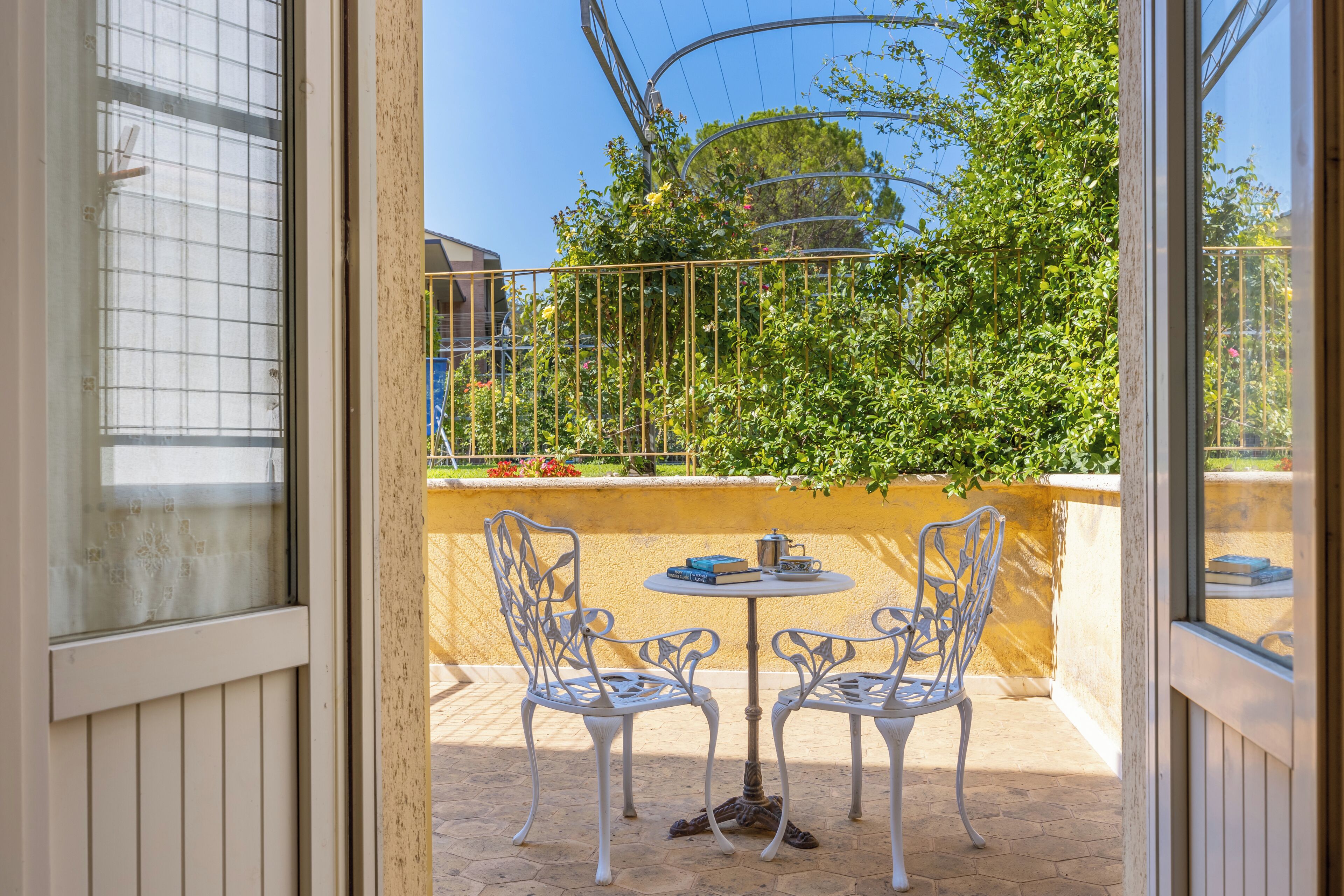 Une terrasse extérieure avec deux chaises en métal blanc et une petite table ronde, un mur jaune et une clôture en métal.