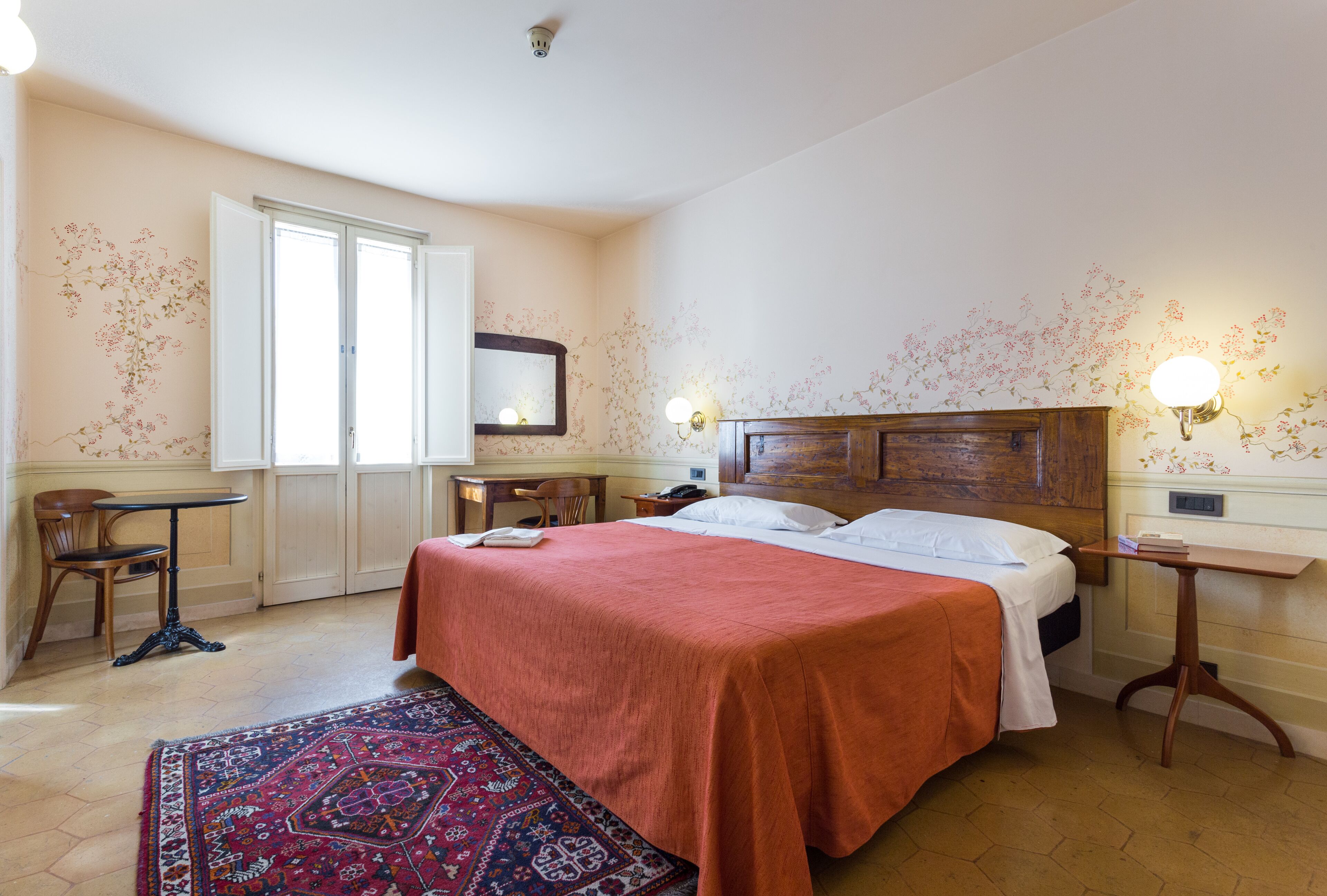 A hotel room with a large bed, a desk, a chair, a small table, and a nightstand.