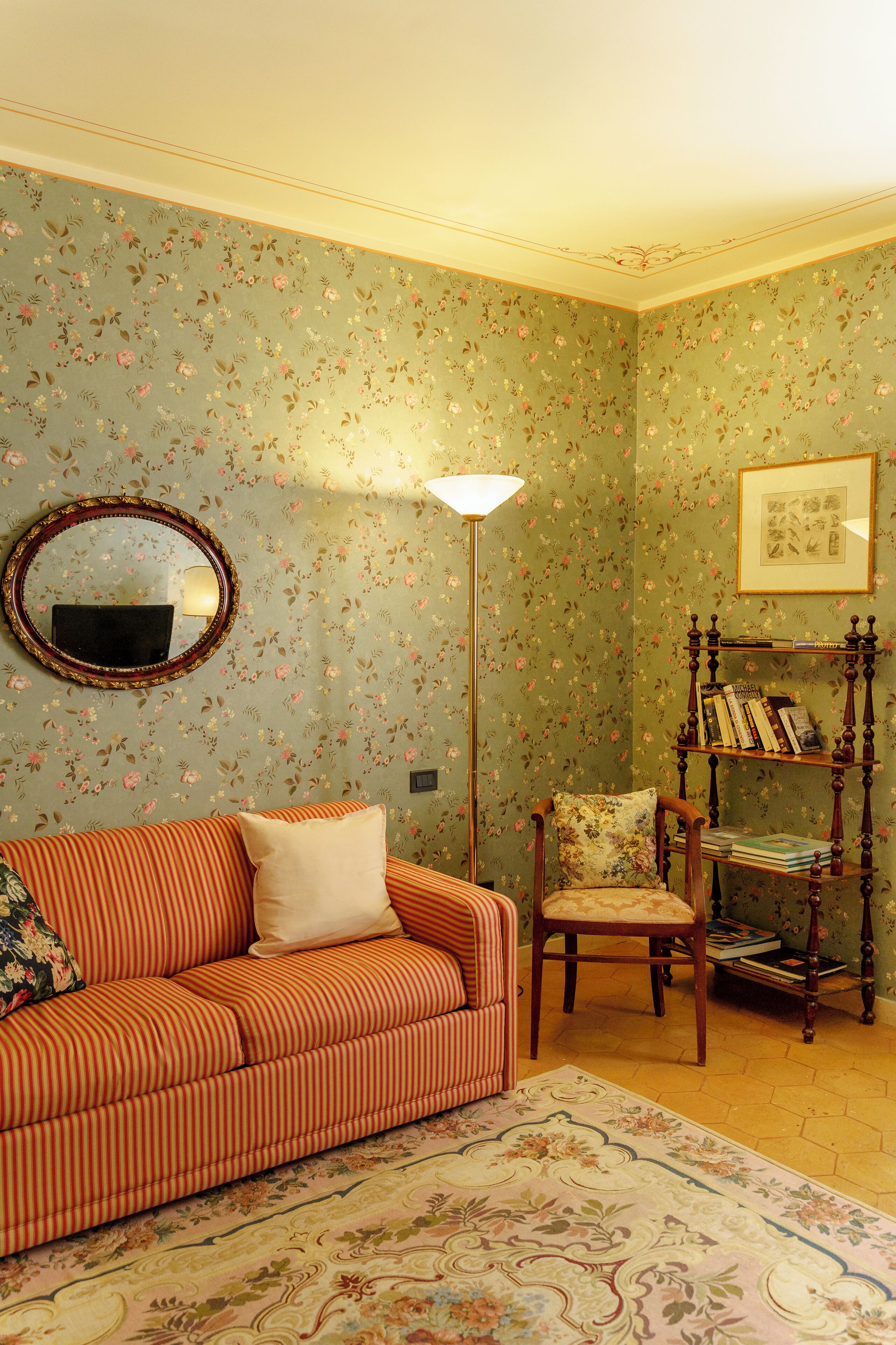 A room with floral wallpaper, a striped sofa, a wooden chair, a floor lamp, and a bookshelf.