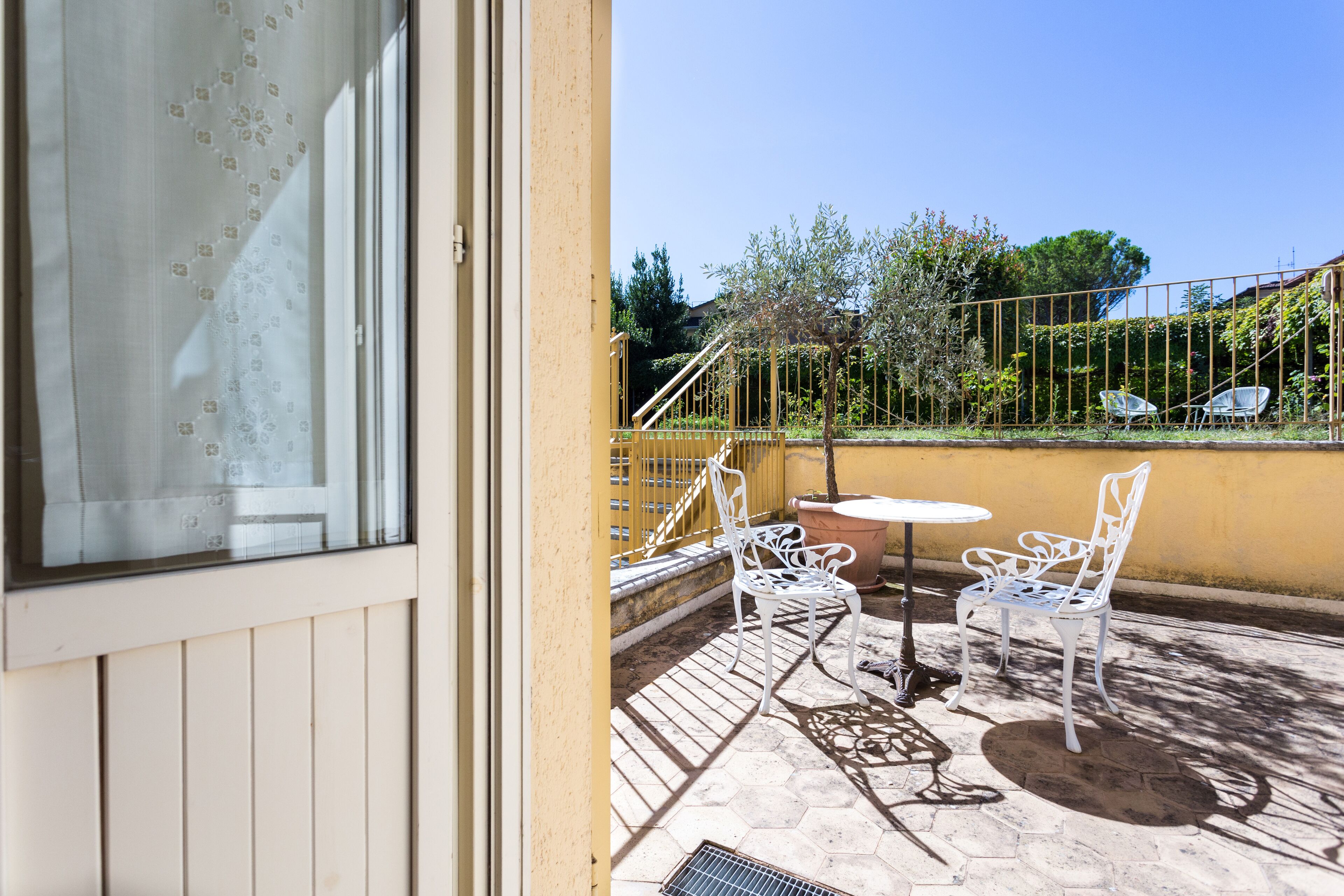 Un balcon avec des chaises en métal blanc et une petite table ronde, un mur jaune et une vue sur un jardin avec des arbres et une clôture.