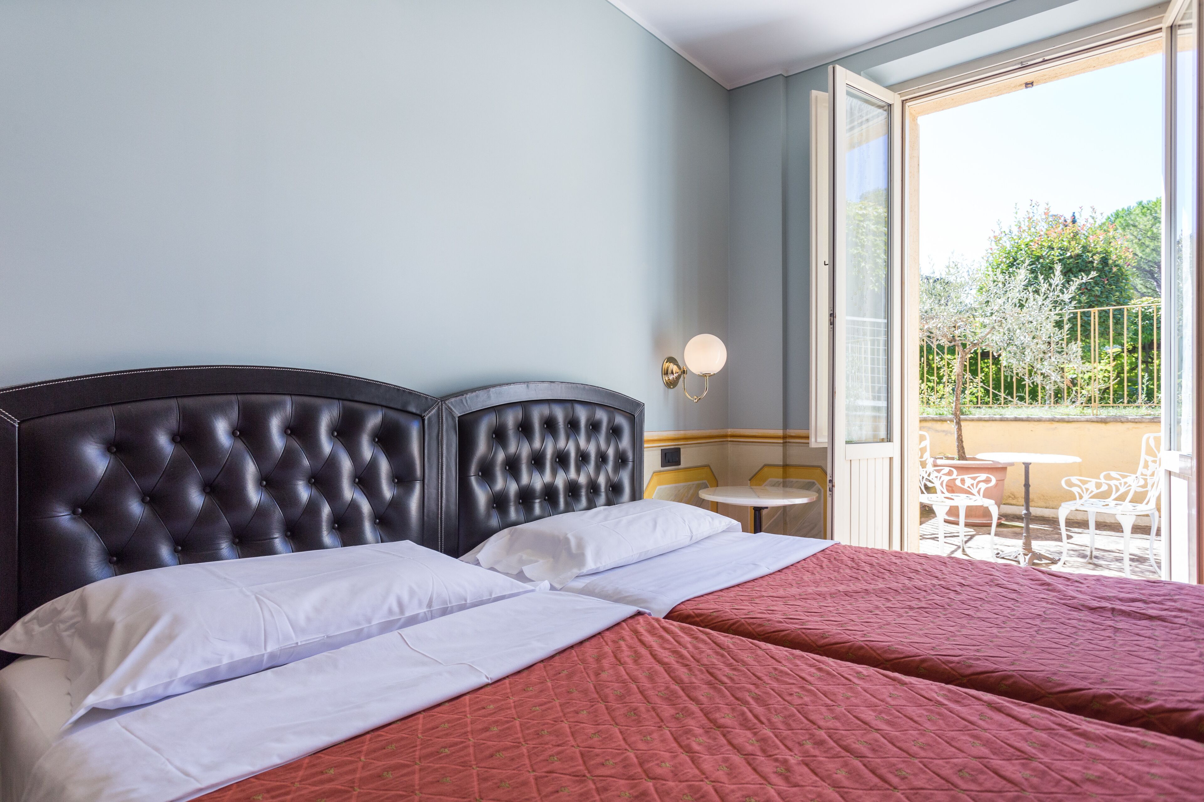A hotel room with a large bed, a balcony with outdoor seating, and a view of greenery.
