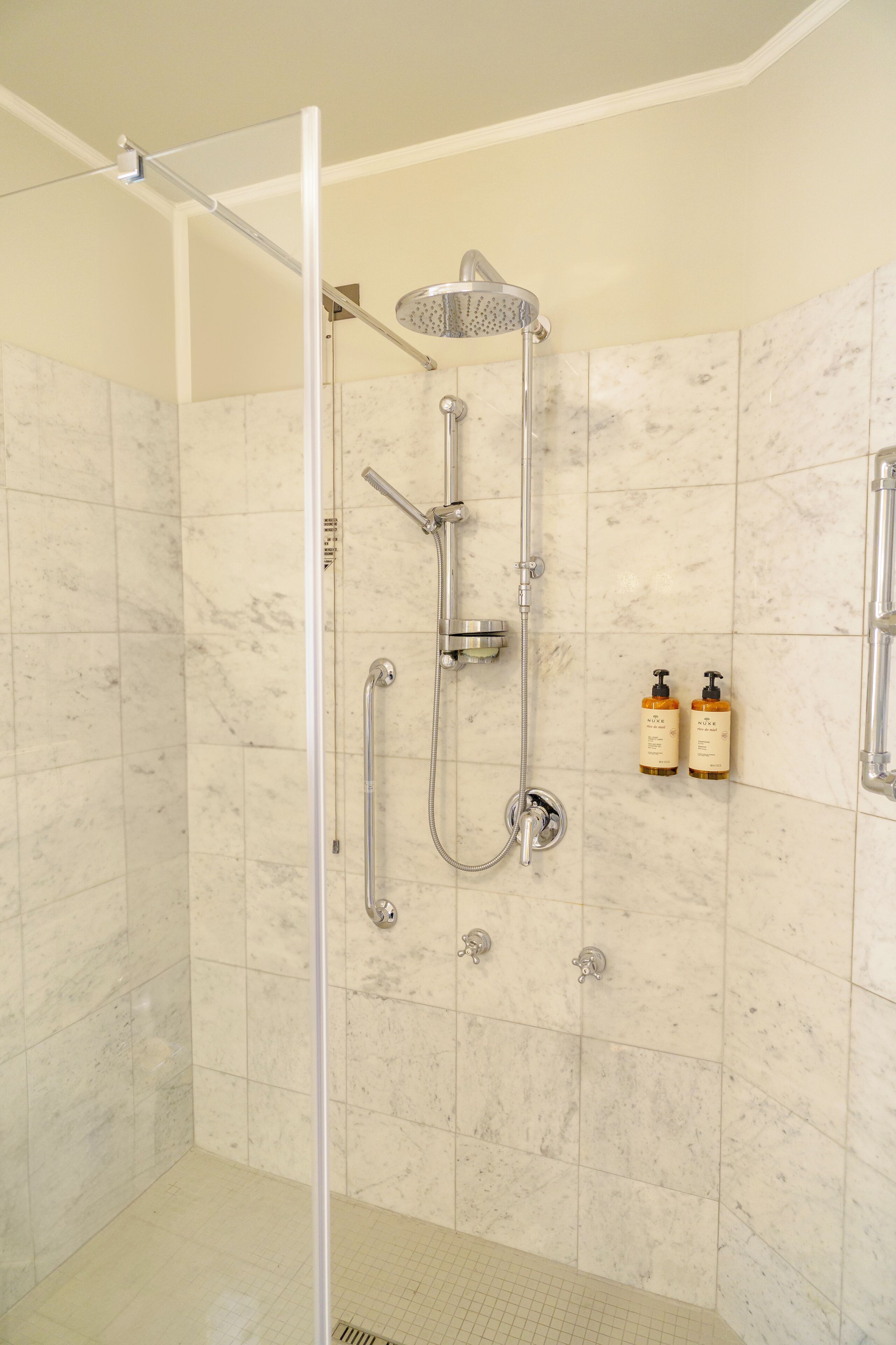 Doccia a filo pavimento con porta in vetro, soffione a mano, rubinetto montato a parete e due flaconi di shampoo sulla parete.