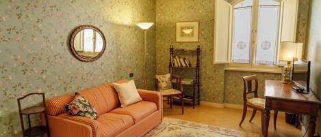 A room with floral wallpaper, a striped sofa, a round mirror, a wooden chair, a small table, and a window with curtains.