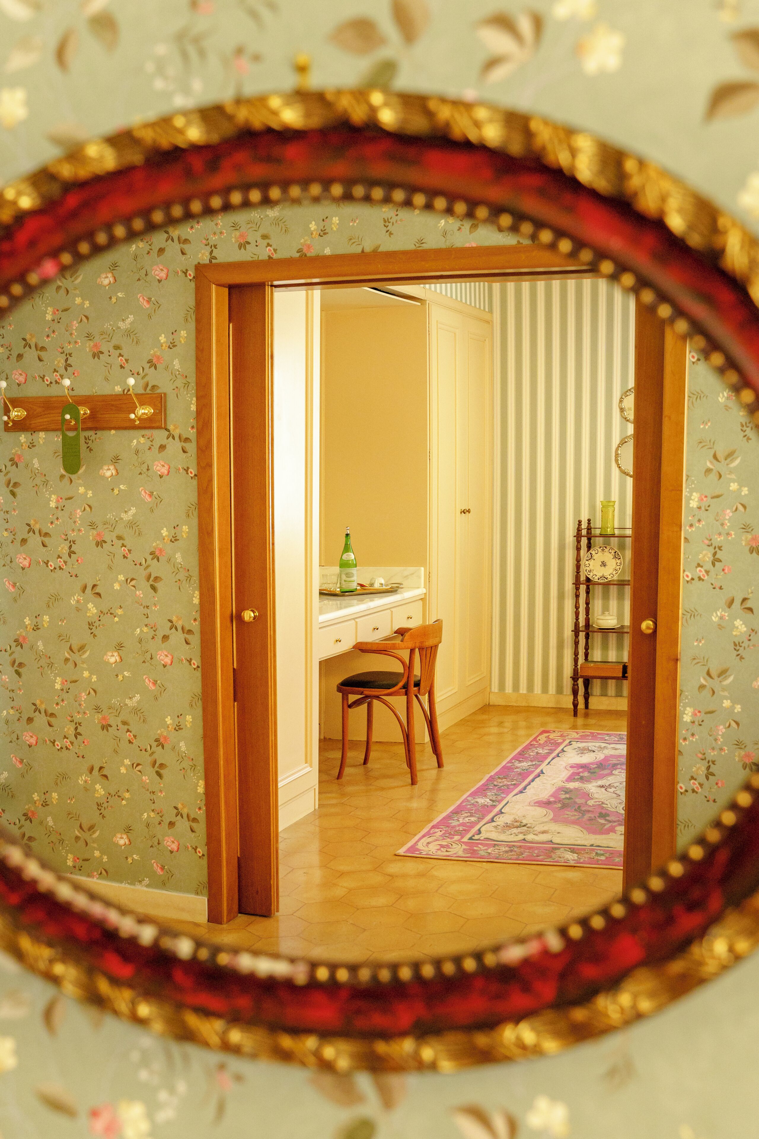 A room with a wooden door, a desk with a chair, a small shelf, and a patterned rug.