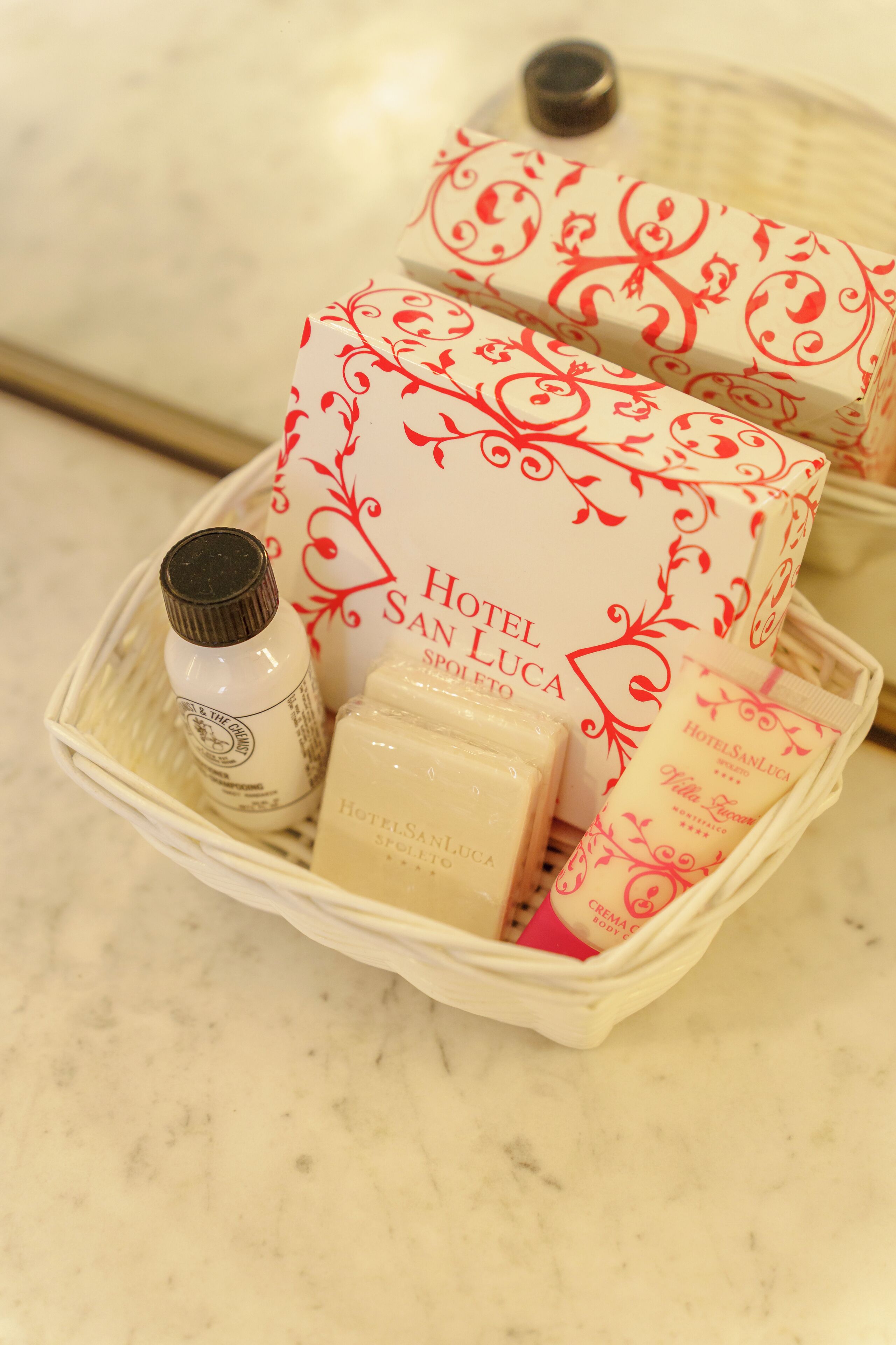 A basket containing Hotel San Luca toiletries, including a soap bar, a cream tube, and a bottle of lotion.