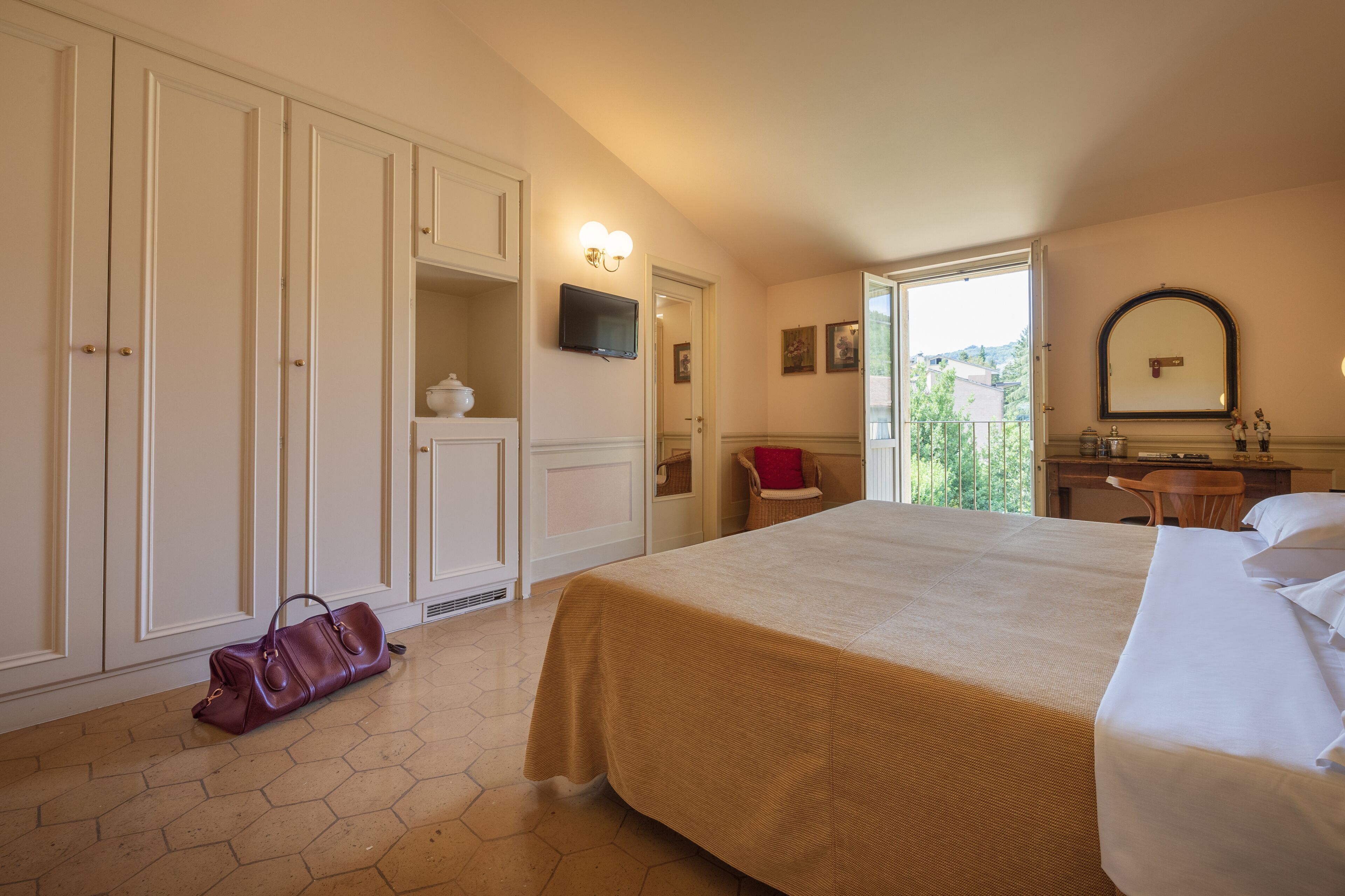 A hotel room with a large bed, a wardrobe, a desk with a chair, a television, and a window with a view of greenery.