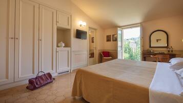 A hotel room with a large bed, a wardrobe, a desk with a chair, a television, and a window with a view of greenery.