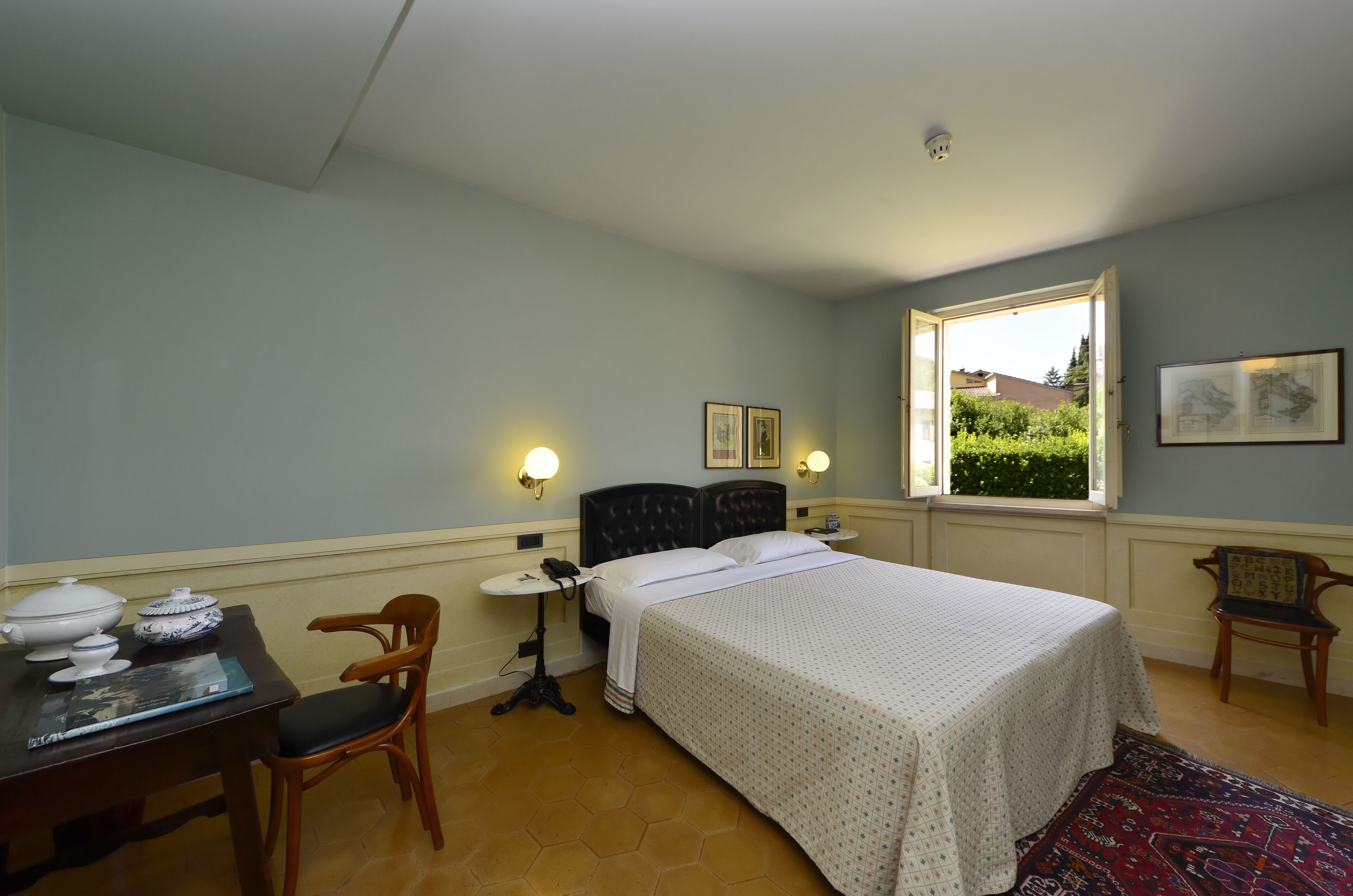 A bedroom with a bed, a desk with a chair, a window with a view, and a wall-mounted light fixture.