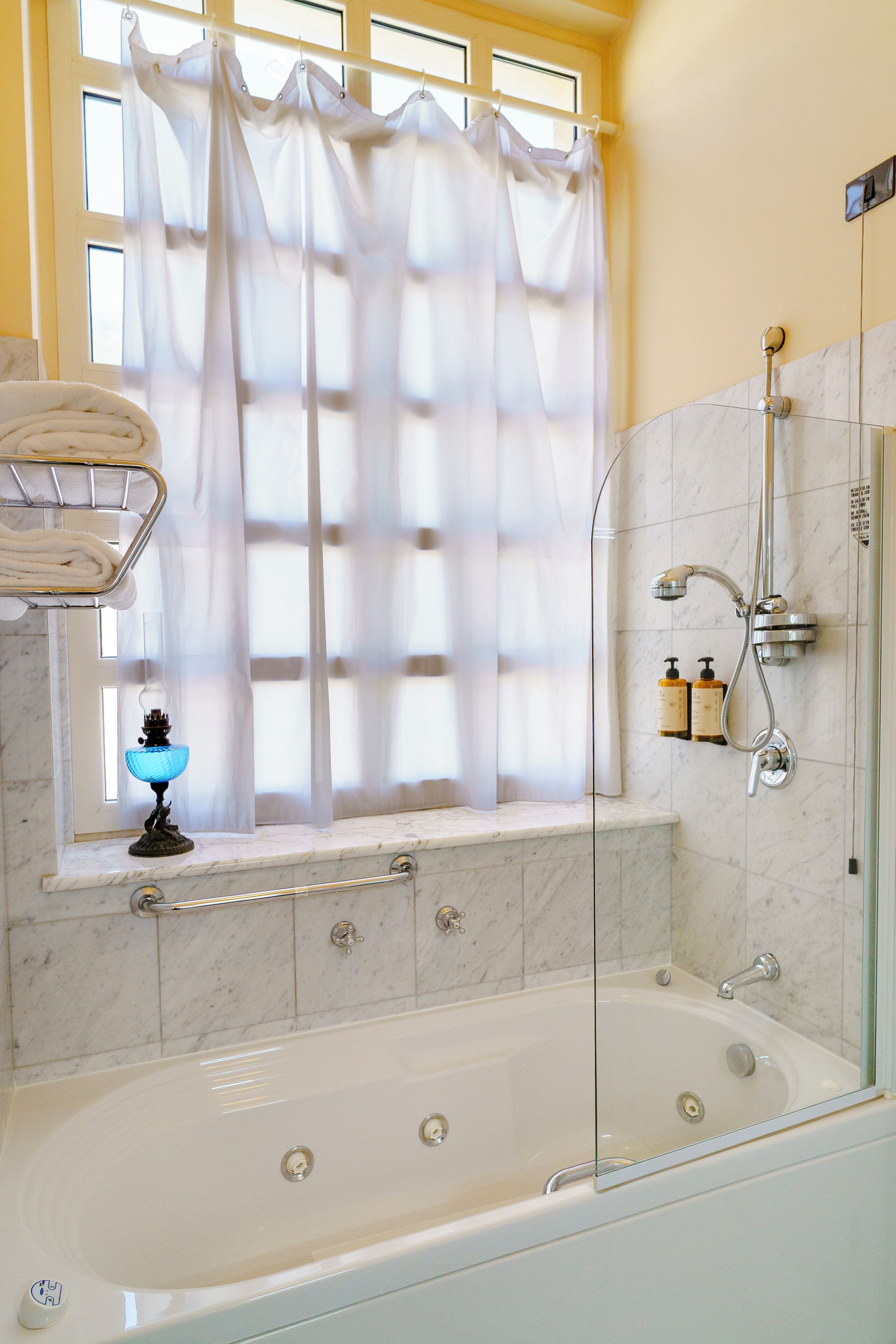 A bathroom with a bathtub, a glass shower enclosure, a window with a sheer curtain, and a towel rack with folded towels.