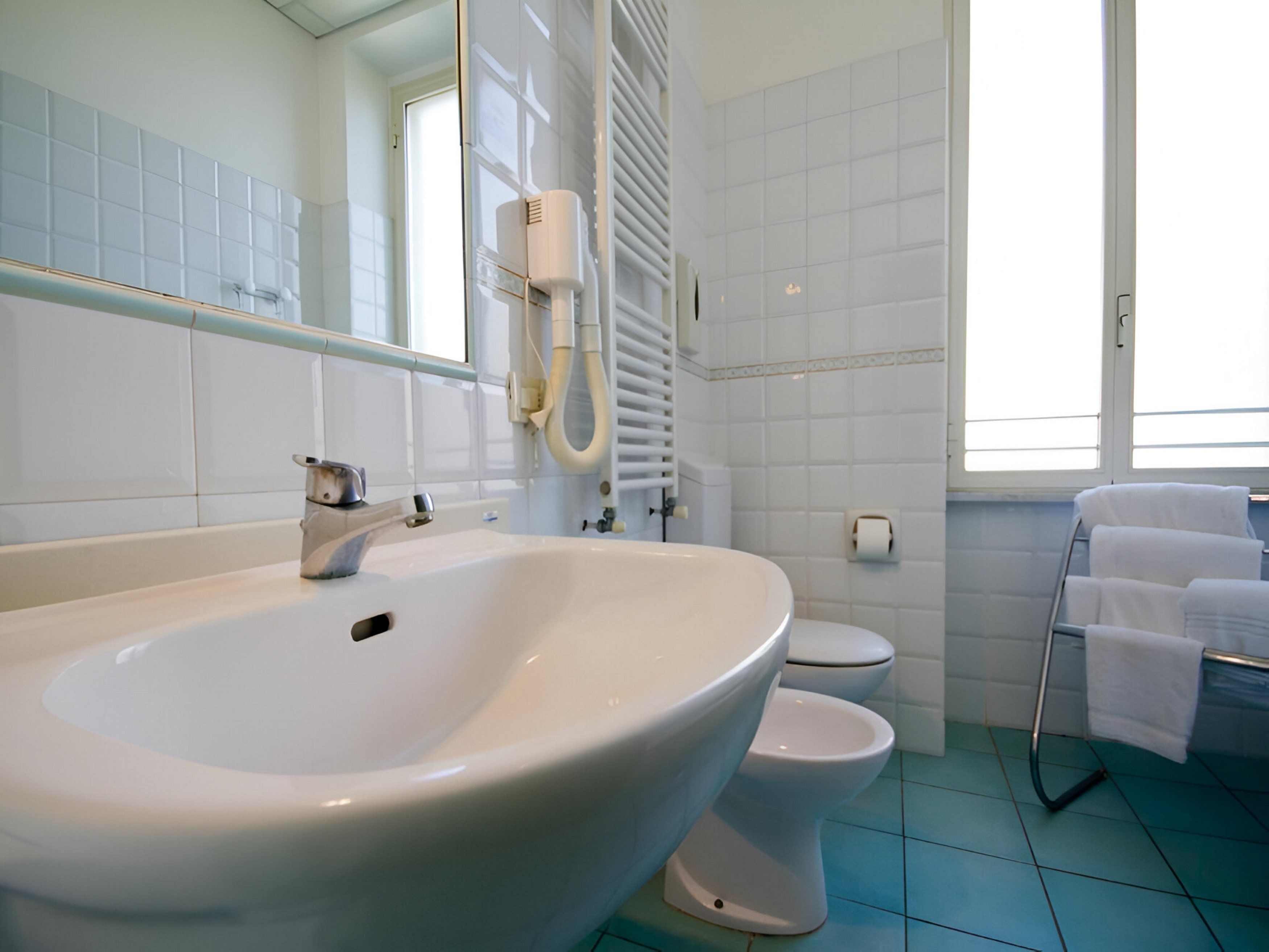 Un baño con un lavamanos grande de color blanco, dos inodoros, un perchero con toallas y una ventana que deja entrar luz natural.