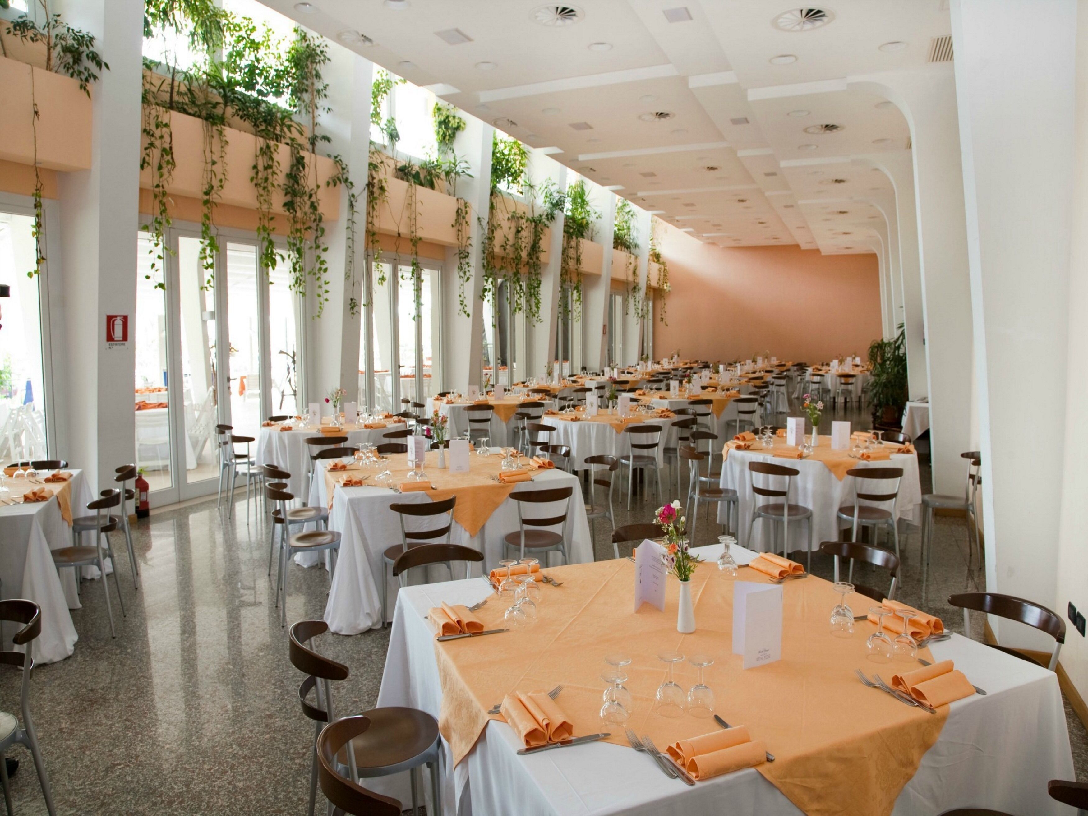 A banquet hall with multiple round tables, each set with white tablecloths, orange napkins, and glassware.