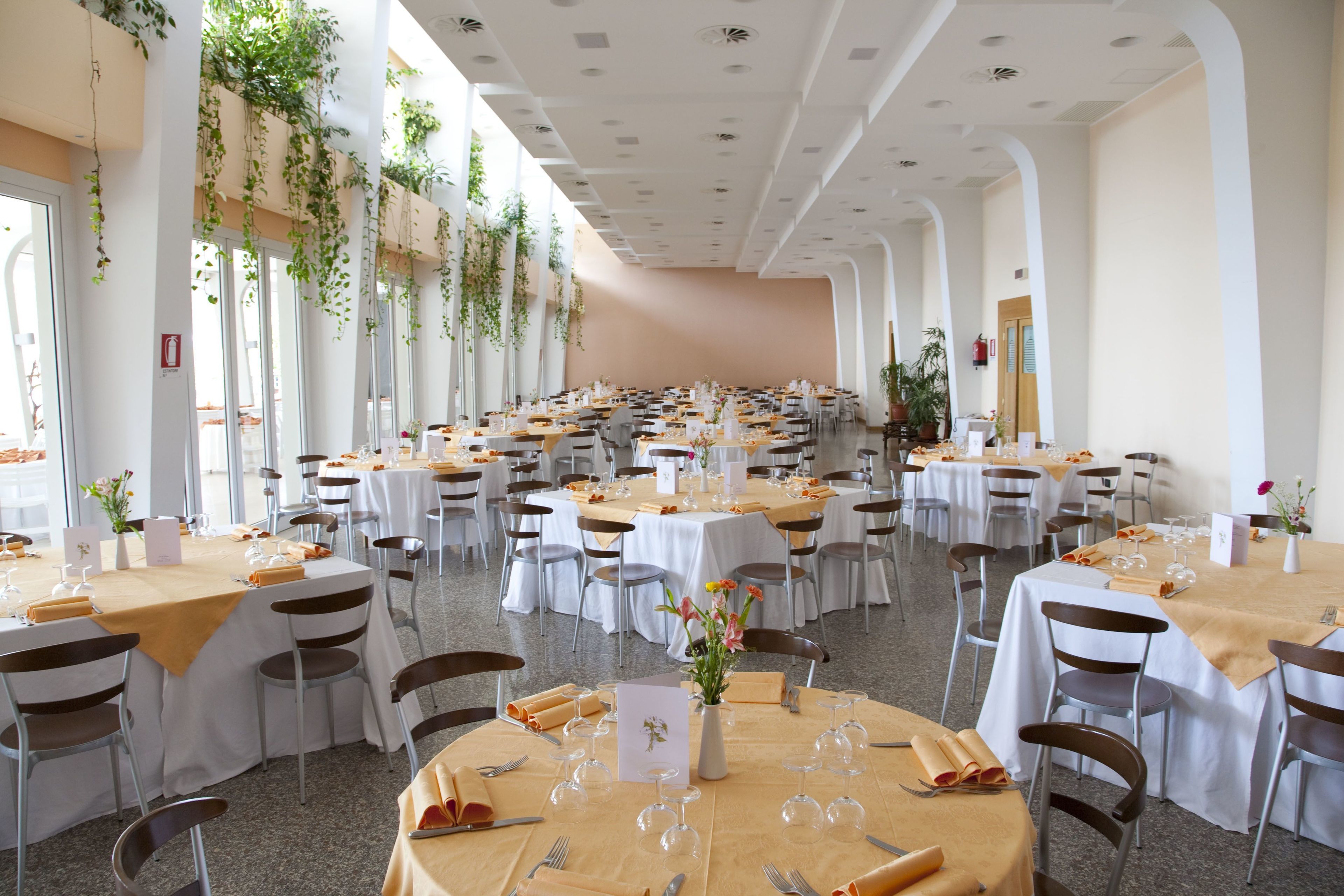 Un amplio salón de banquetes con varias mesas redondas, cada una con mantel blanco, cubertería y copas de vino.