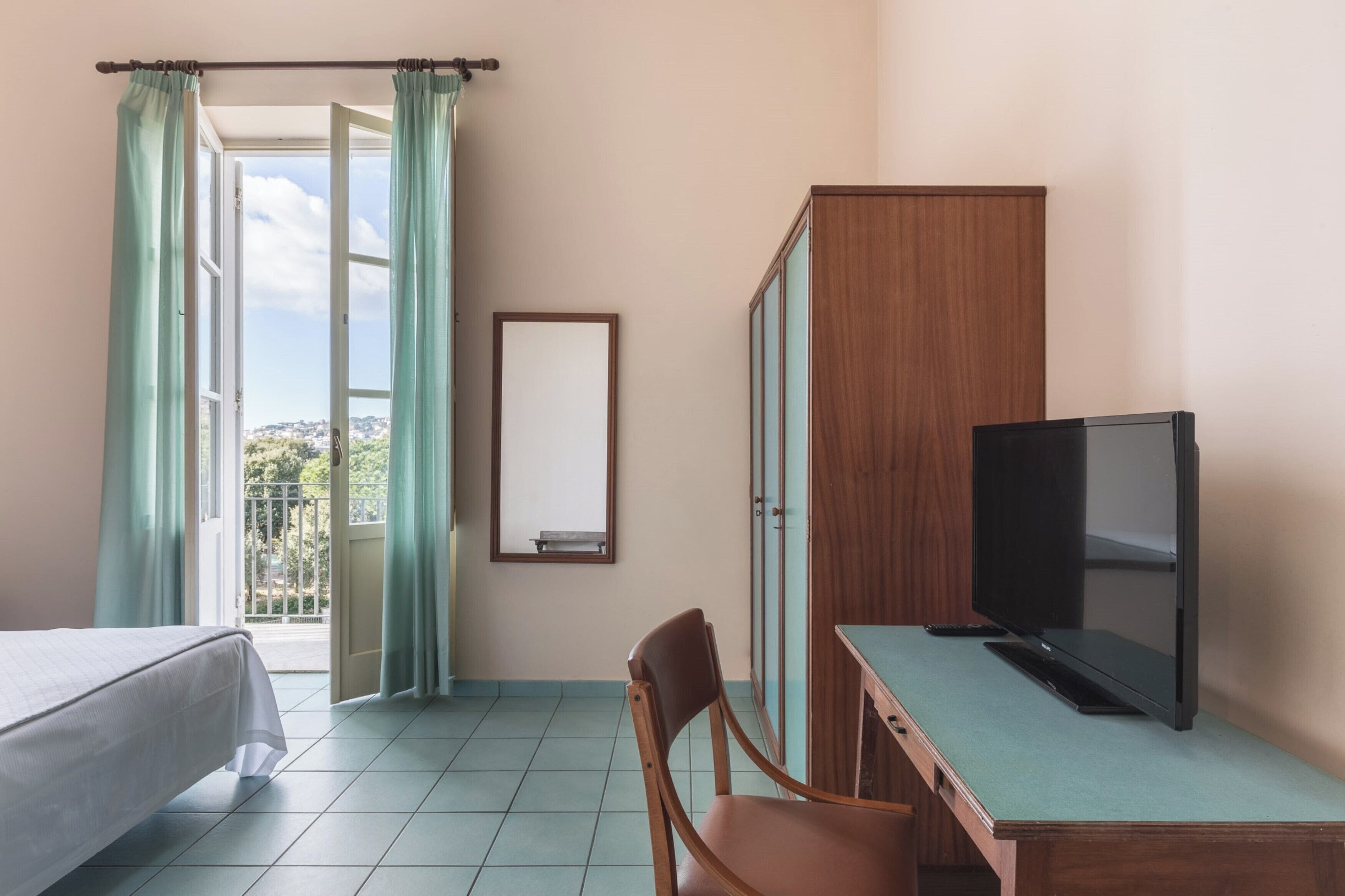 A hotel room with a bed, a desk with a TV, a chair, and a wardrobe.
