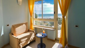 A room with a beige sofa, a small round table with a drink, and a window with yellow curtains. - Hotel Serapo (Gaeta)