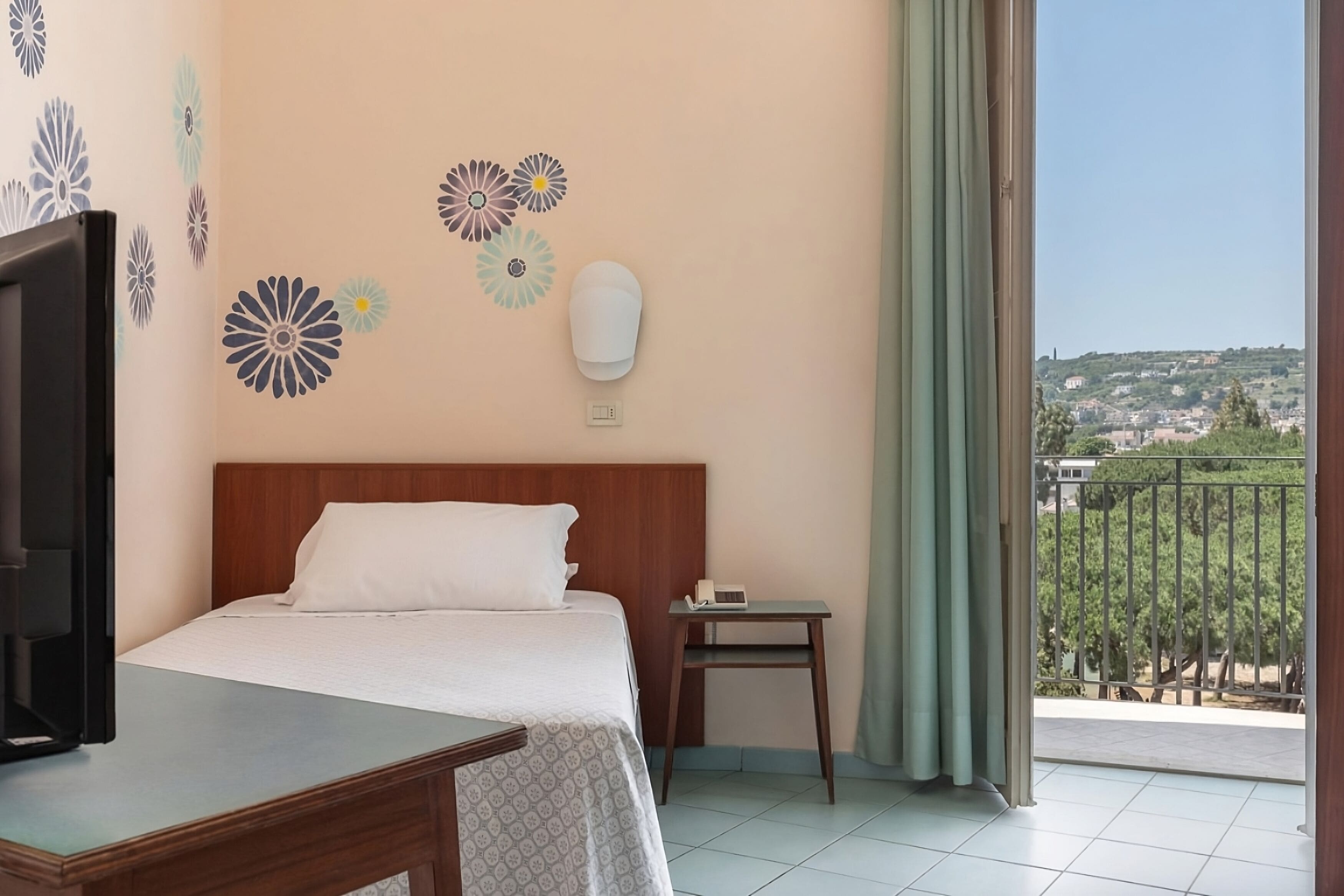 Una camera d'albergo con un letto, un comodino con il telefono, adesivi decorativi al muro e vista su un balcone con ringhiera.
