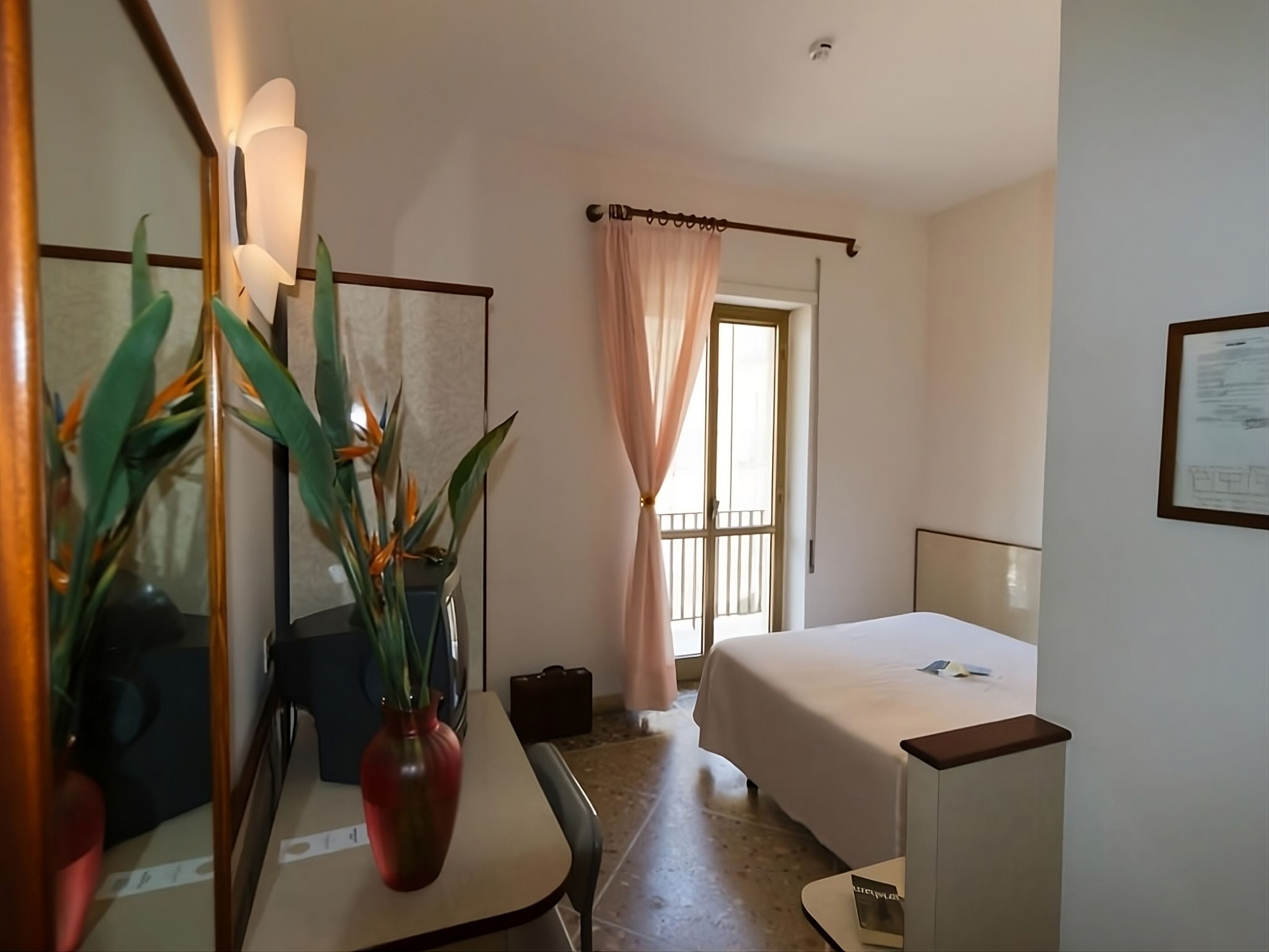 A hotel room with a bed, a desk with a vase of flowers, a mirror, and a window with curtains.