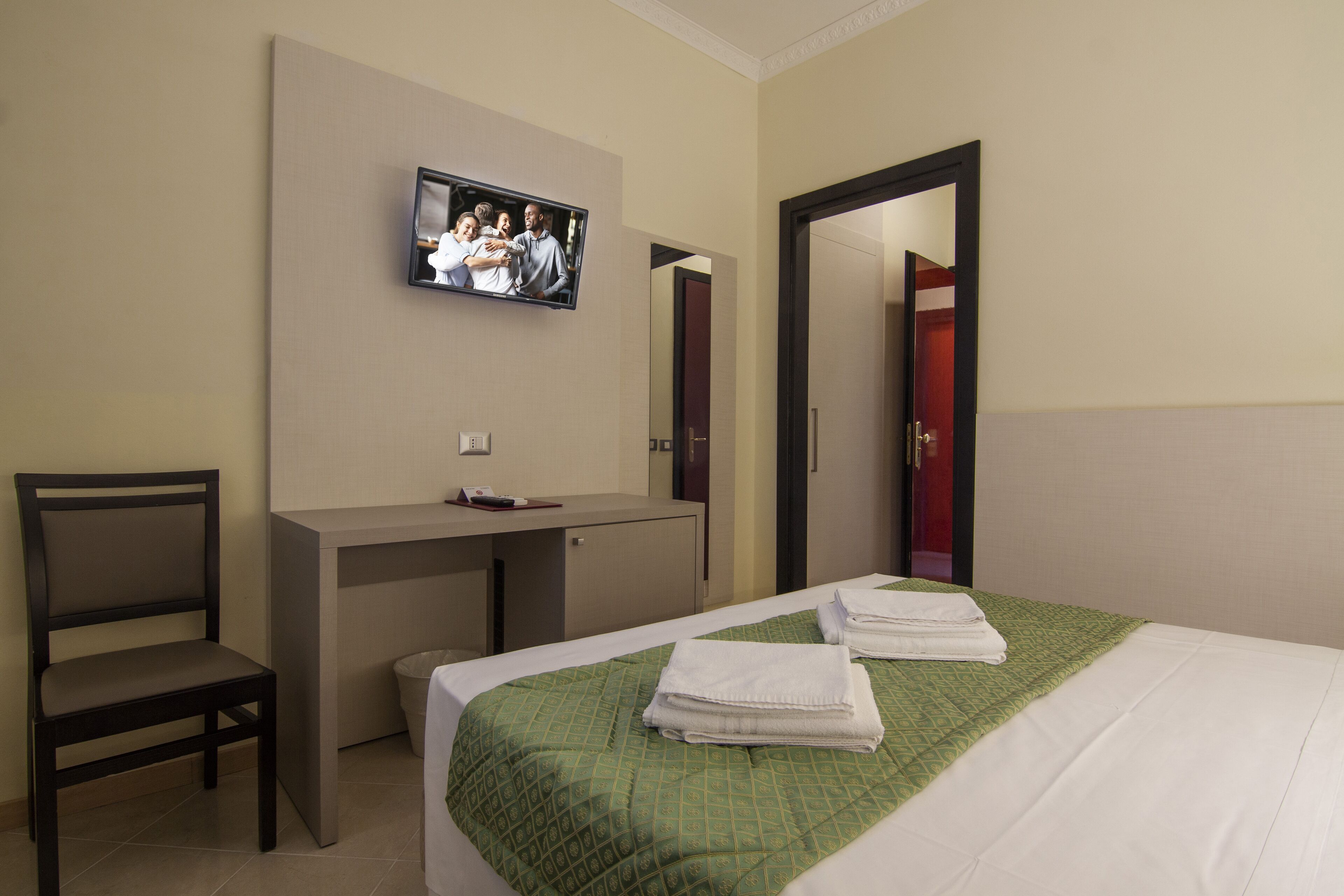 Une chambre d’hôtel comprenant un lit, un bureau, une chaise, un téléviseur fixé au mur et une porte donnant sur une autre pièce.
