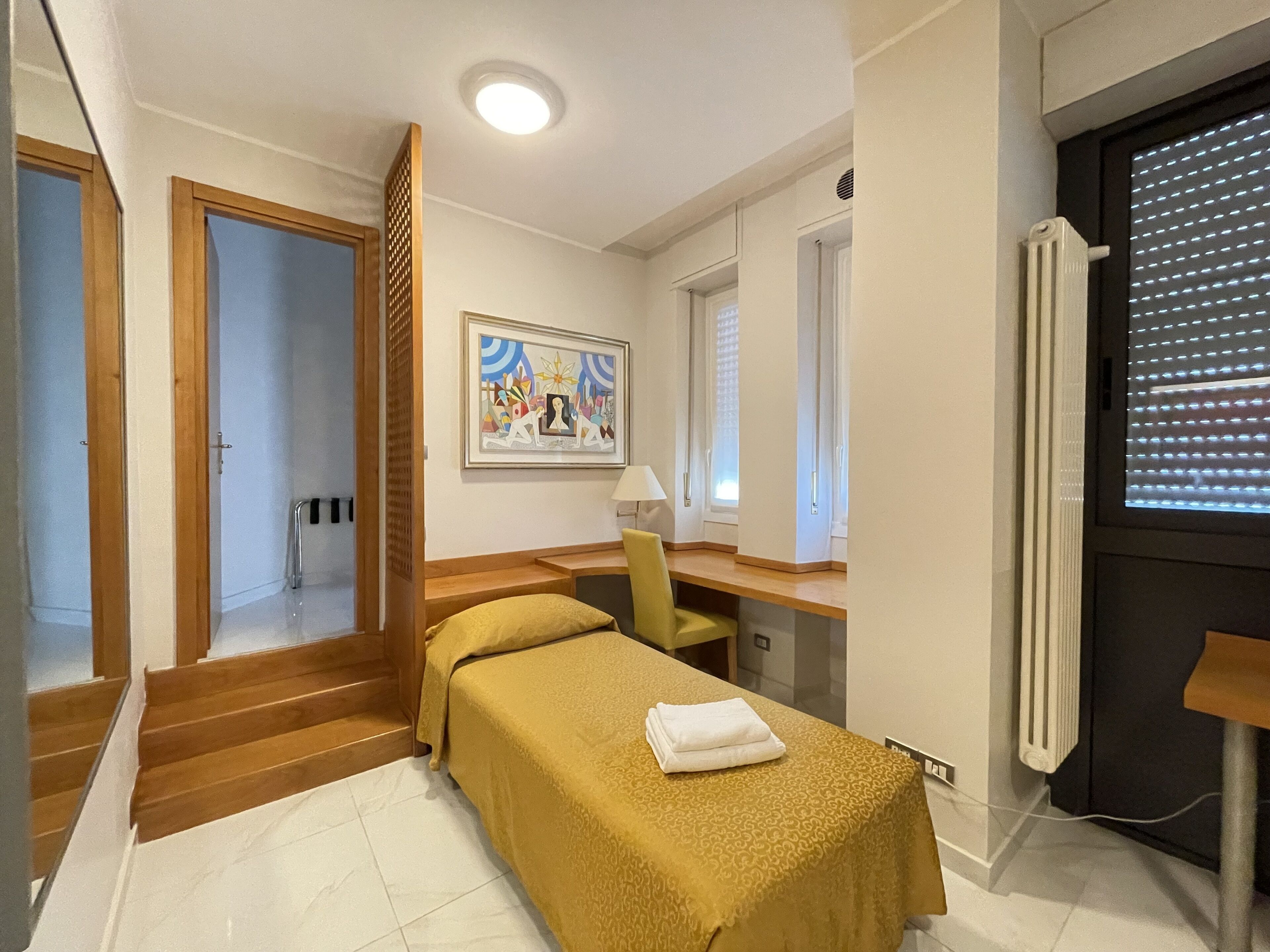 A hotel room with a bed, a desk, a chair, a painting on the wall, and a window with blinds.