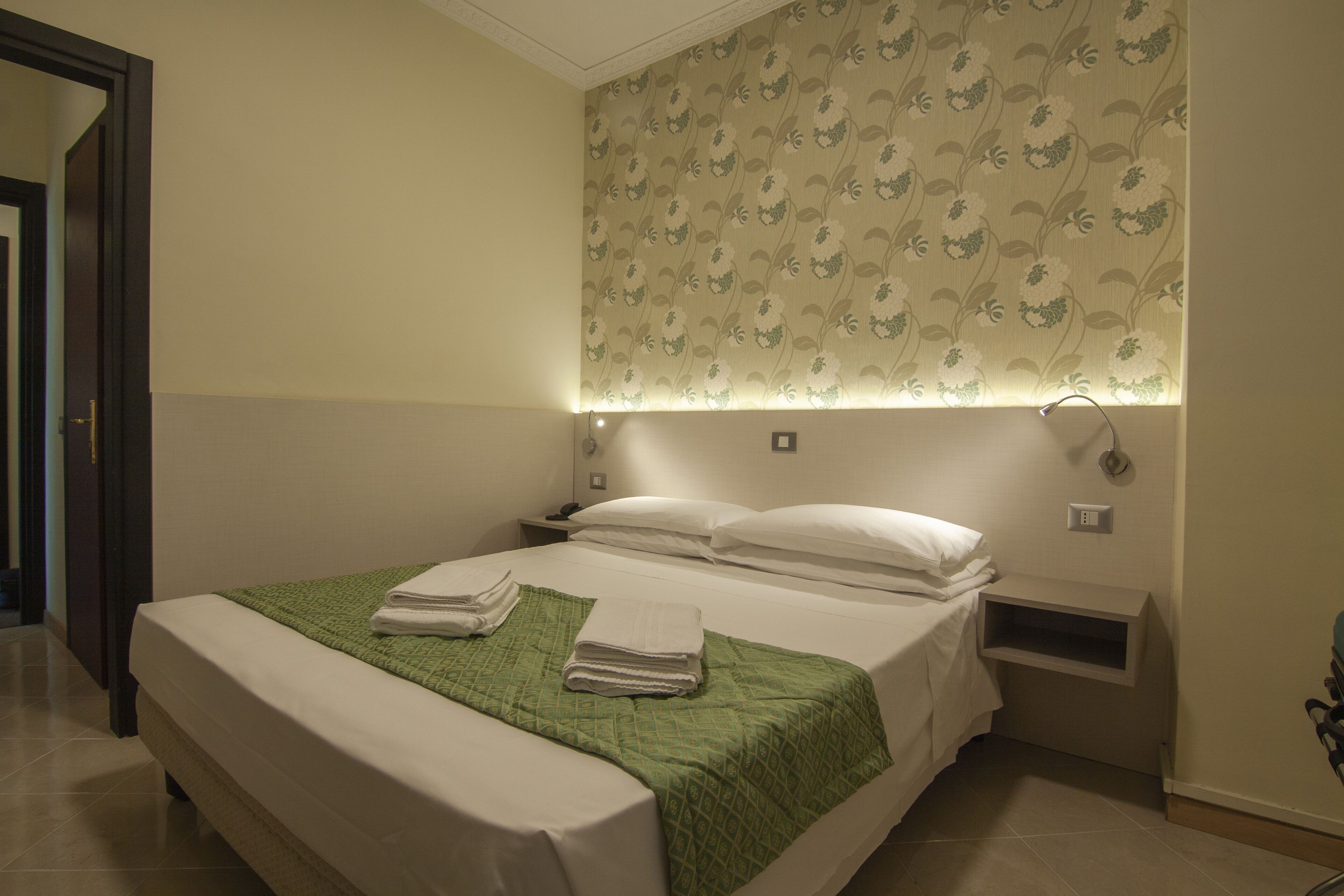 A neatly made bed with a green blanket and white pillows, a bedside table with a lamp, and a wall with floral wallpaper.