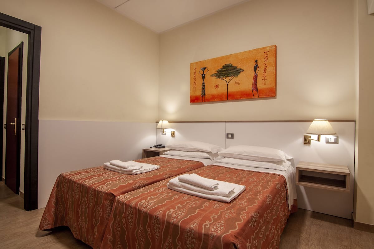 Ett hotellrum med två sängar, var och en med ett mönstrat överkast och prydligt vikta handdukar. Det finns sänglampor och en tavla på väggen ovanför sängarna.
