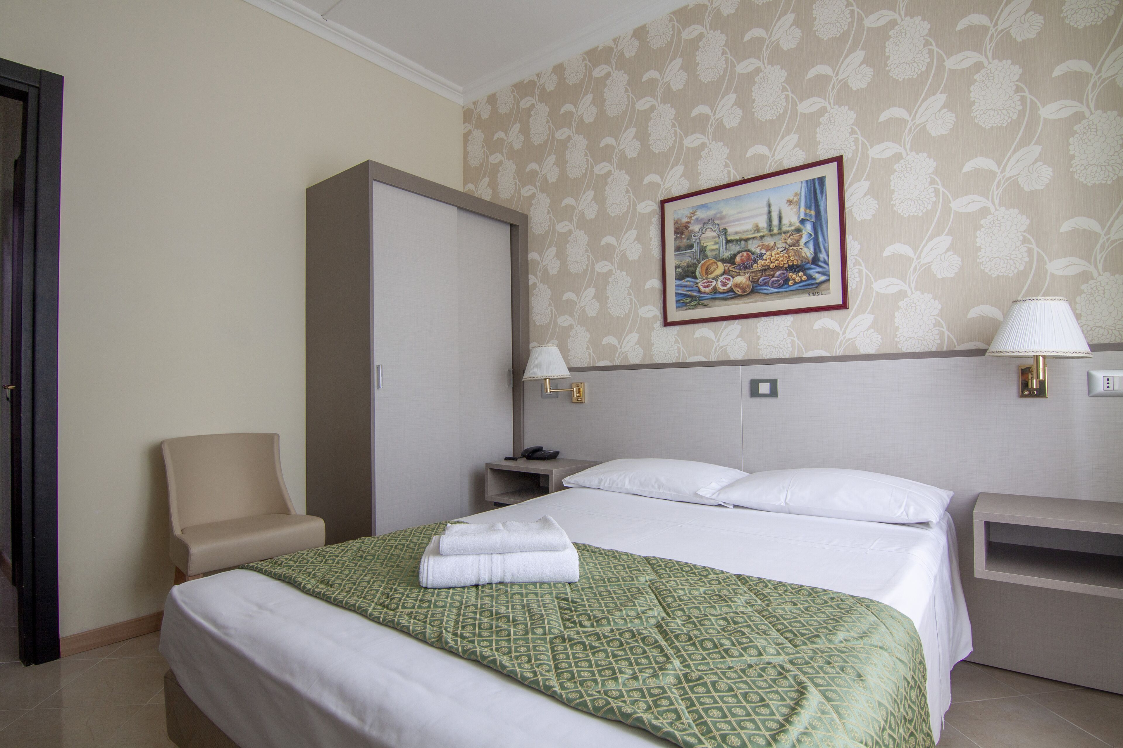 Una habitación de hotel con una cama, una silla, una mesita de noche, una lámpara y un cuadro en la pared.
