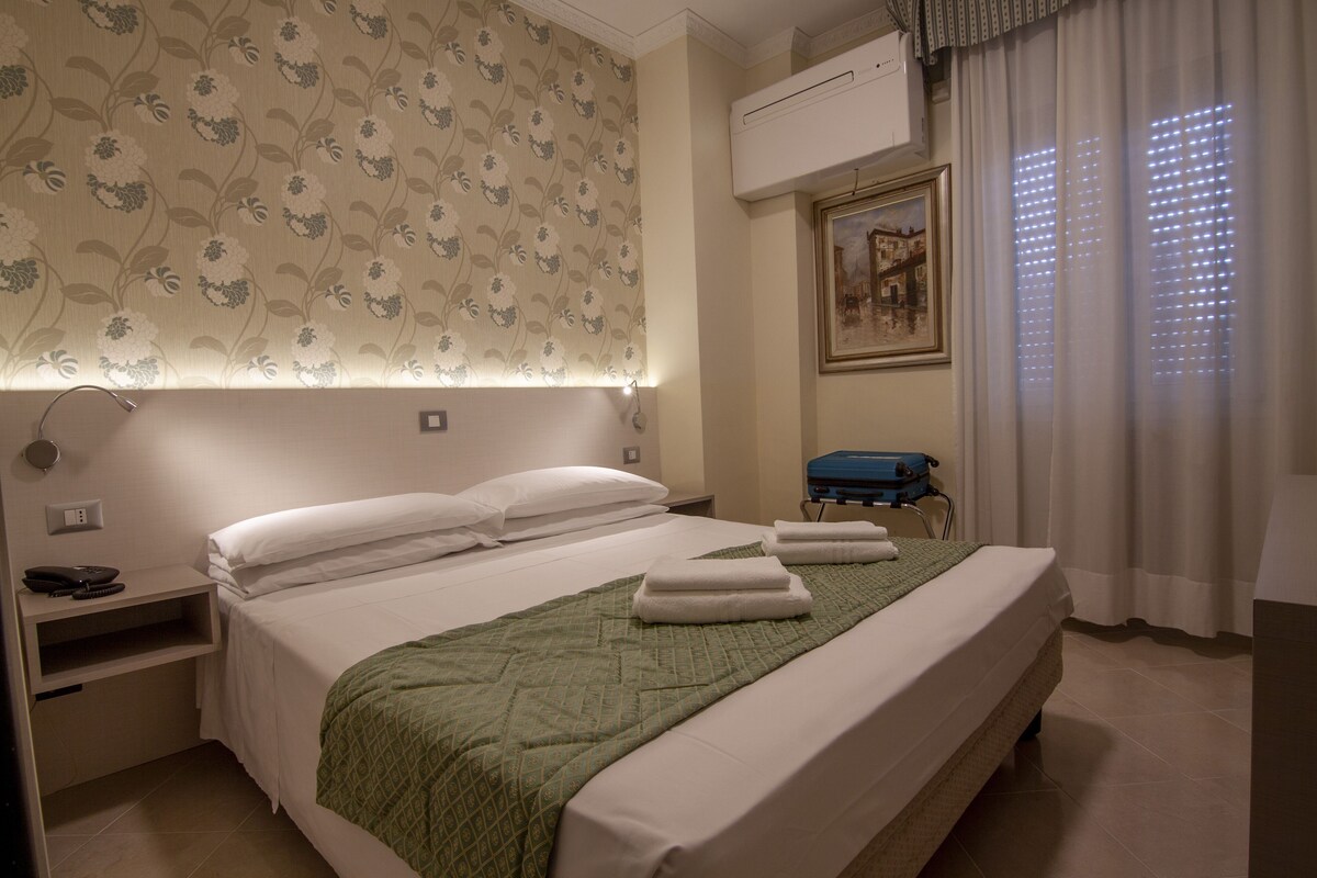 A hotel room with a bed, bedside table, lamp, air conditioning unit, and a window with curtains.