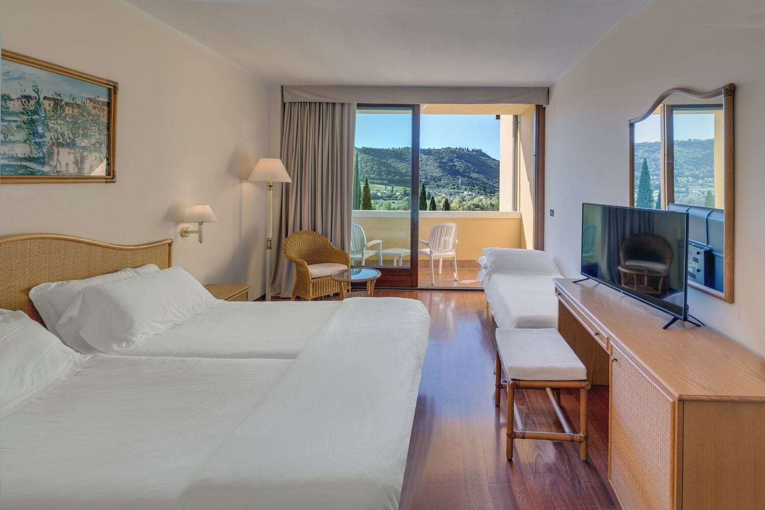 A hotel room with two beds, a TV, a balcony with a view of the hills, and a wicker chair.