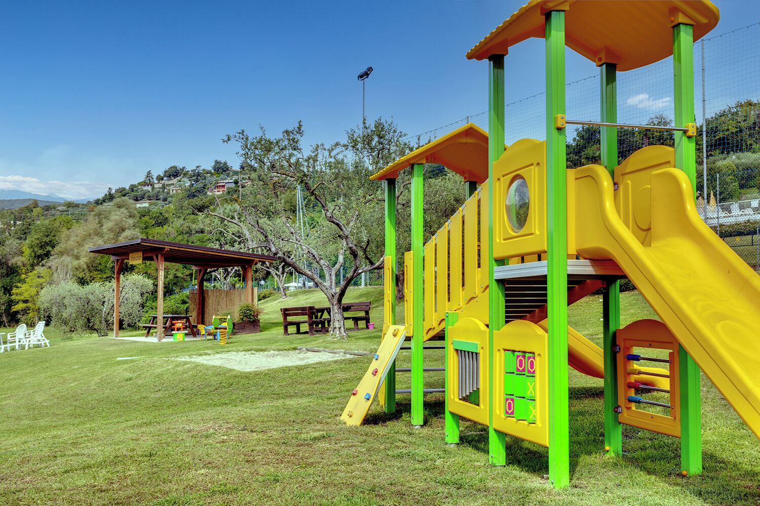 Une aire de jeux avec un toboggan et des structures d’escalade, des tables de pique-nique et un espace de repos abrité dans un parc.