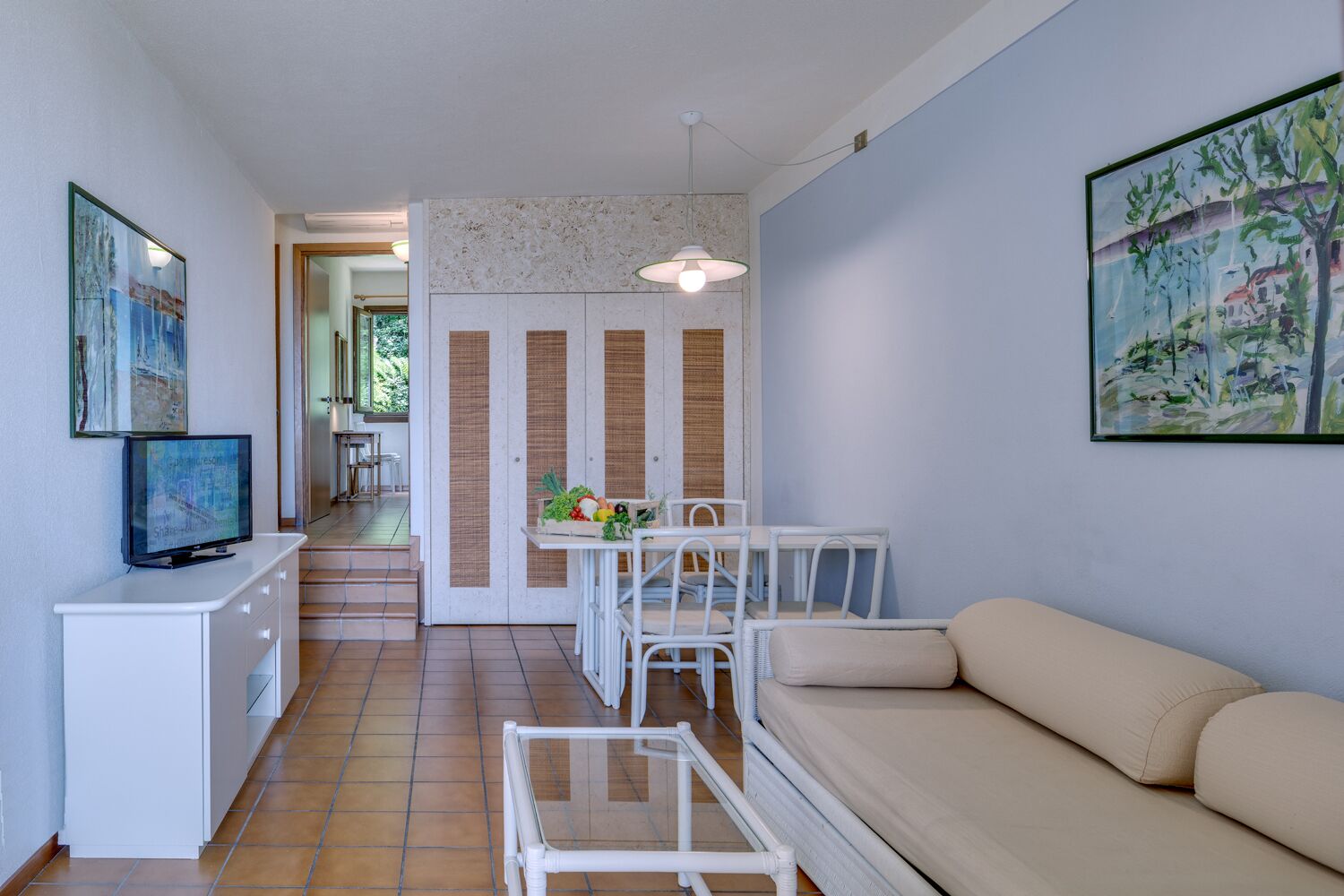 A modern living room with a sofa, a dining table with chairs, a TV on a cabinet, and artwork on the walls.