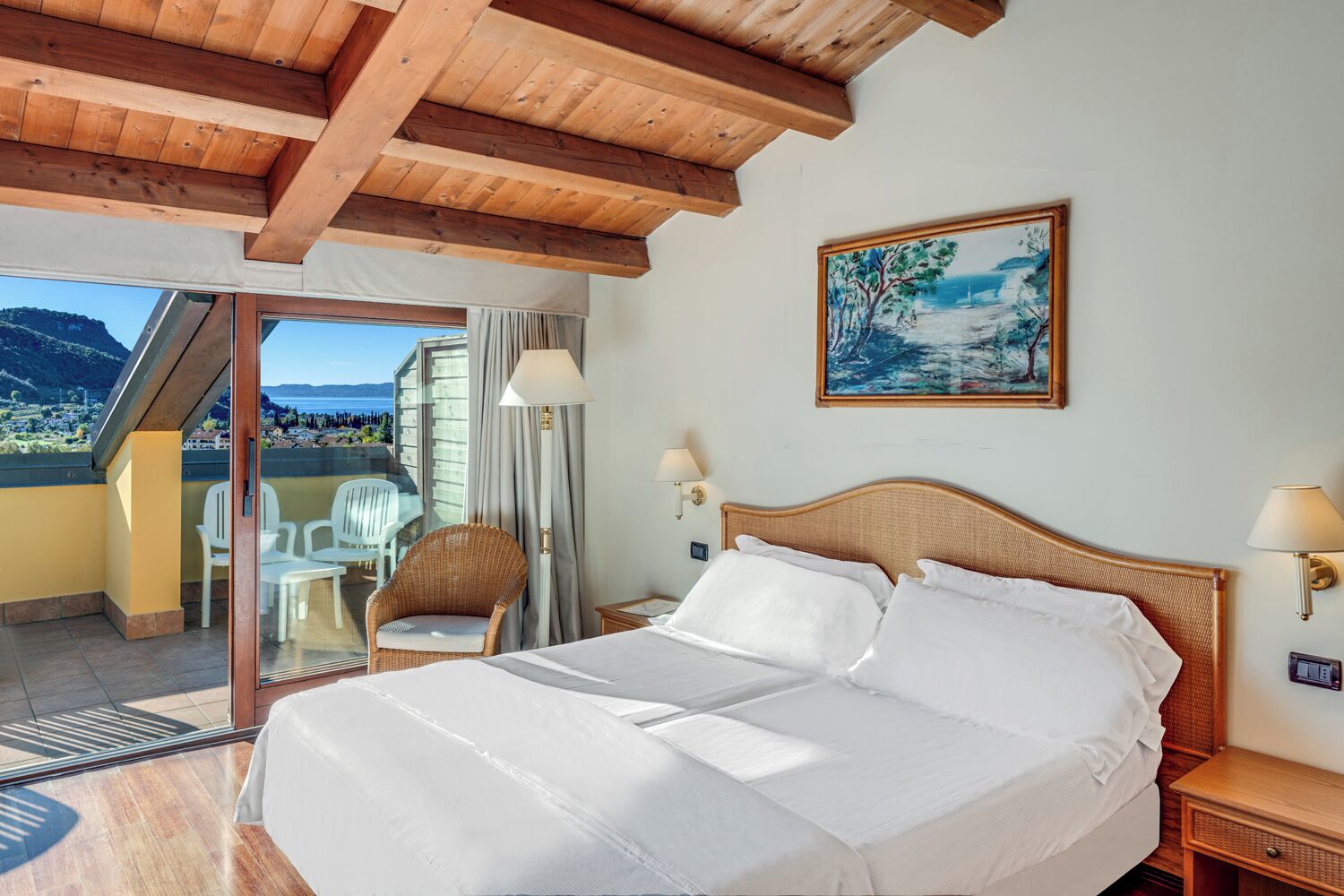A hotel room with a large bed, a balcony with chairs, and a painting on the wall.