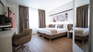 A modern hotel room with a bed, a desk with a computer, a chair, and a large mirror.