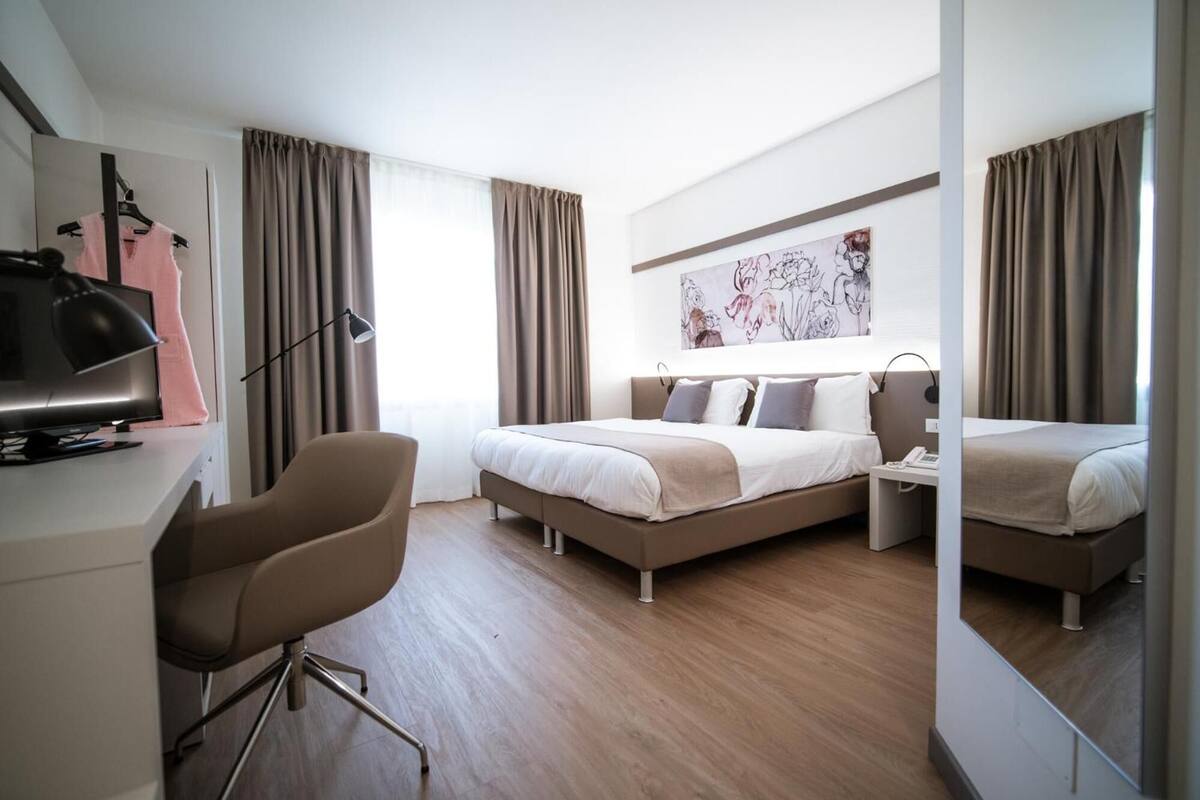 A modern hotel room with a bed, a desk with a computer, a chair, and a large mirror.